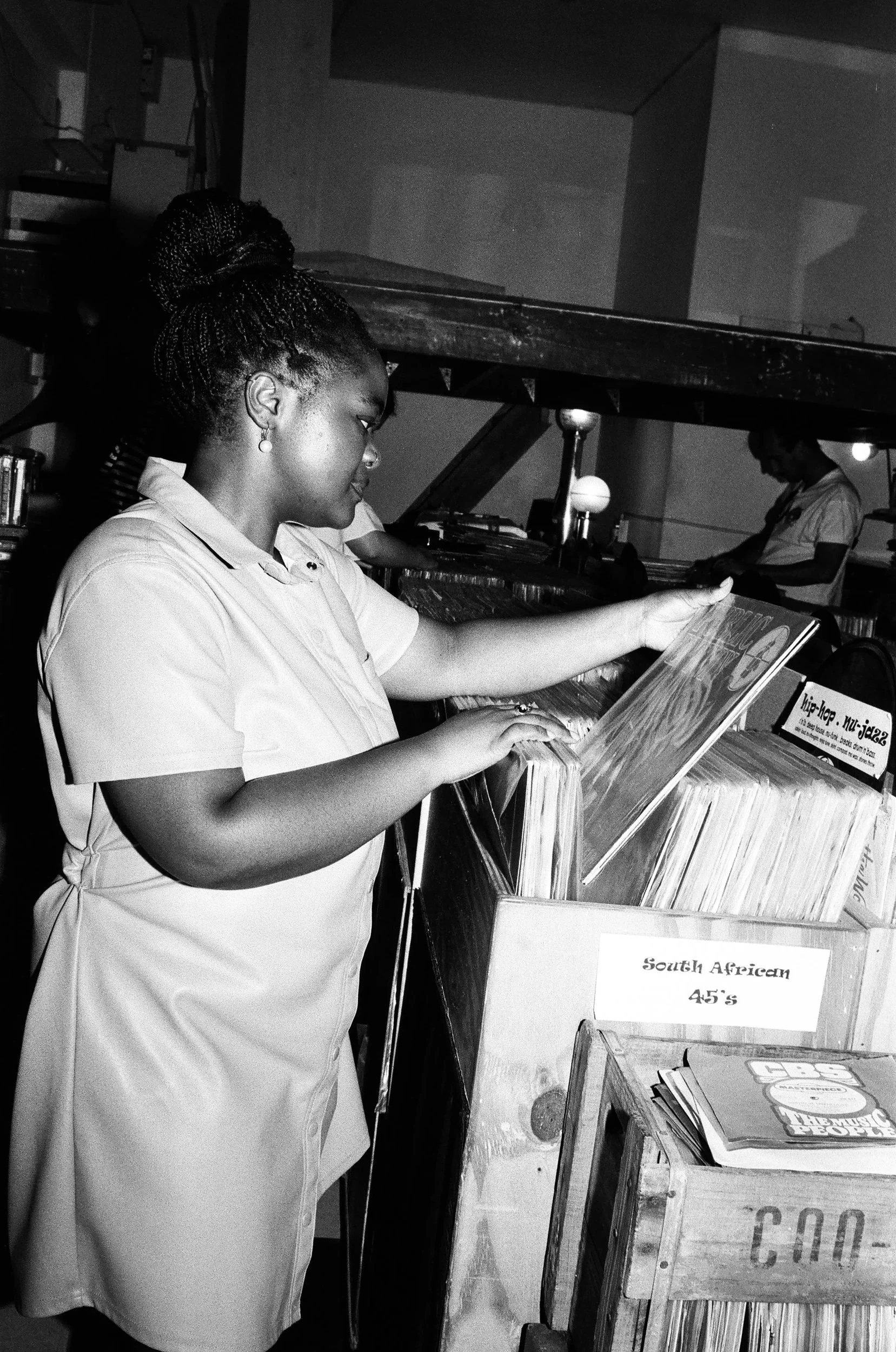 Zizipho Bam browsing vinyl records in a black and white photo, with a sign that reads "South African 45's" in a record store or music shop.