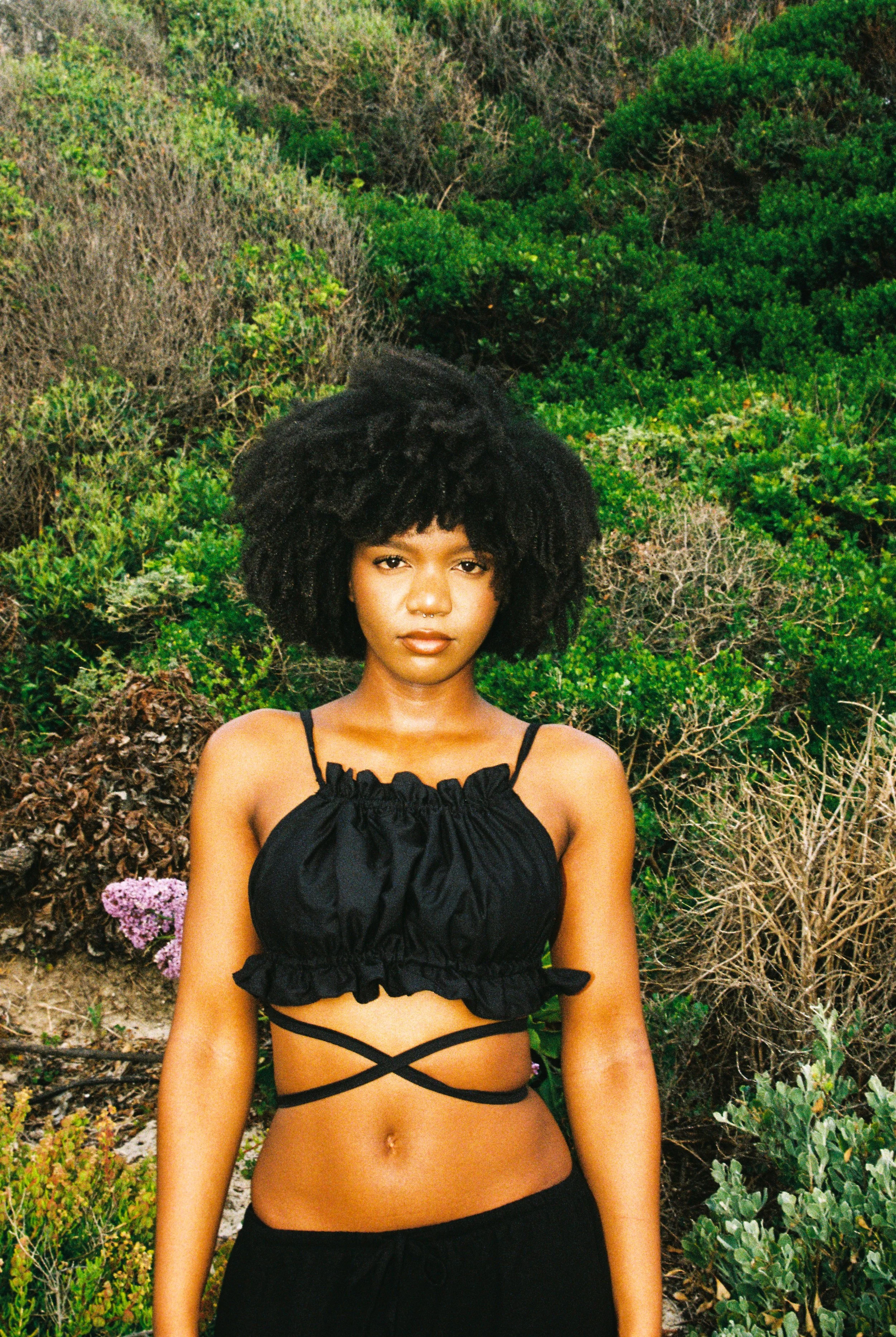 Young woman with curly black hair wearing a black crop top with spaghetti straps and crisscross straps around her waist, standing in front of green bushes and plants.