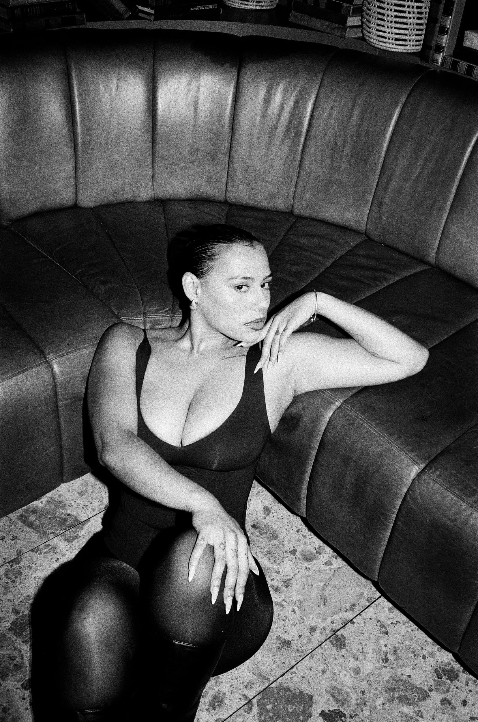 Kimberleigh Venty sitting on the floor, leaning against a curved leather couch with her arm resting on it. She is wearing a sleeveless, low-cut black dress and jewelry, looking at the camera.