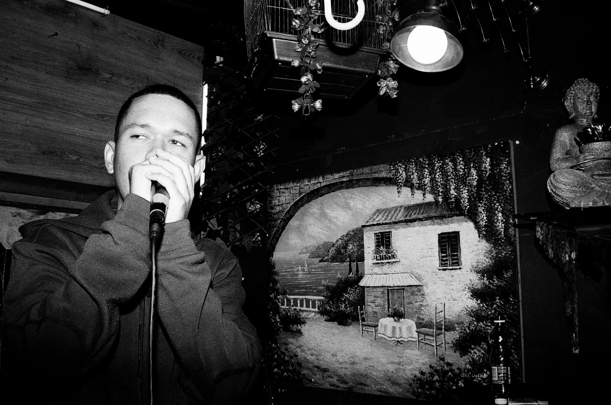 Jarry Pitboi holding a microphone, singing or speaking, in front of a mural of a lakeside house and scenery in a dimly lit room.