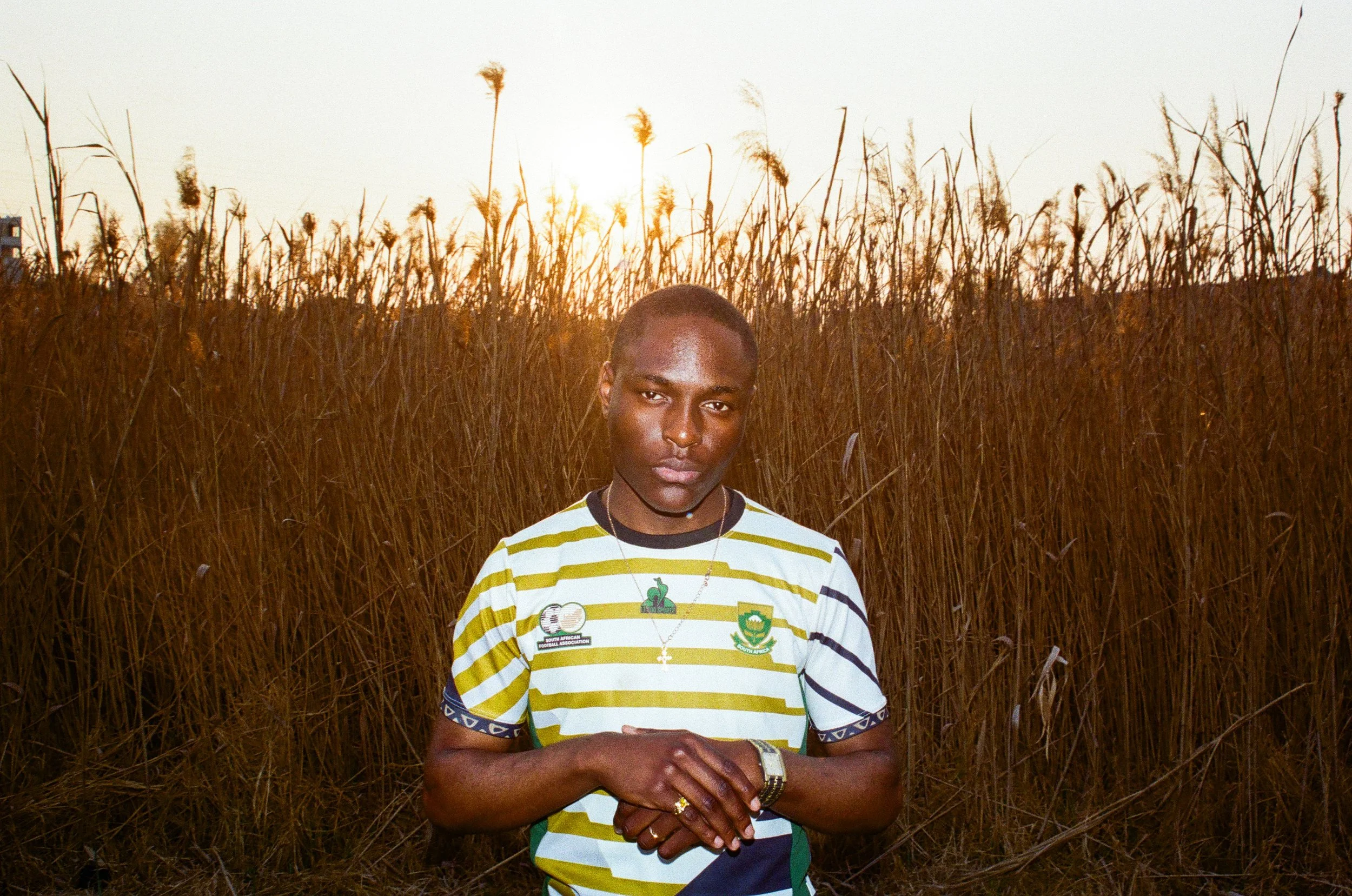 Joshua Futura standing in a field of tall, dry grass at sunset, wearing a yellow and white striped shirt with patches and necklaces.