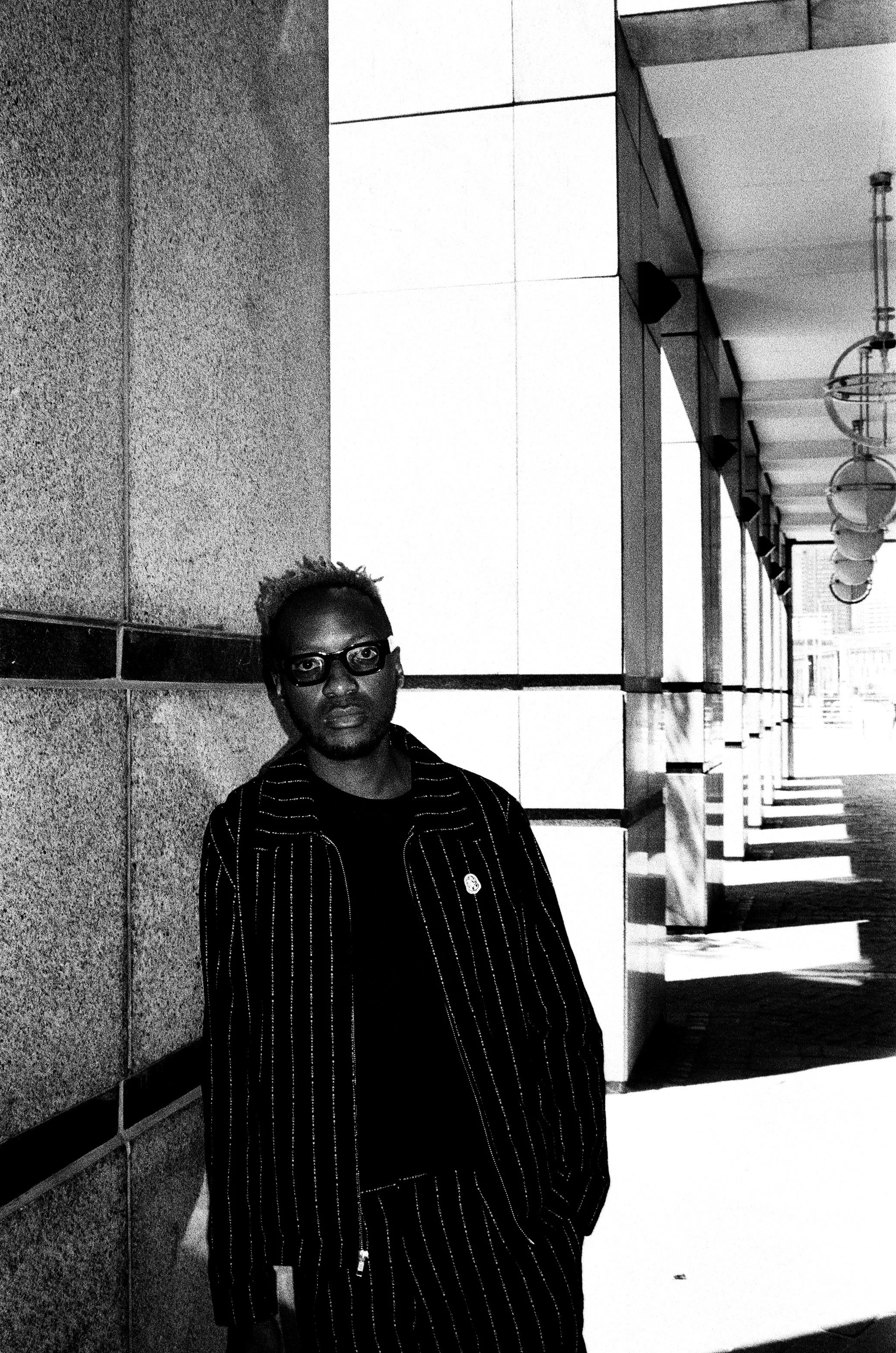 A man with glasses wearing a striped suit standing against a wall outside a building with a row of hanging lamps and cast shadows on the ground.