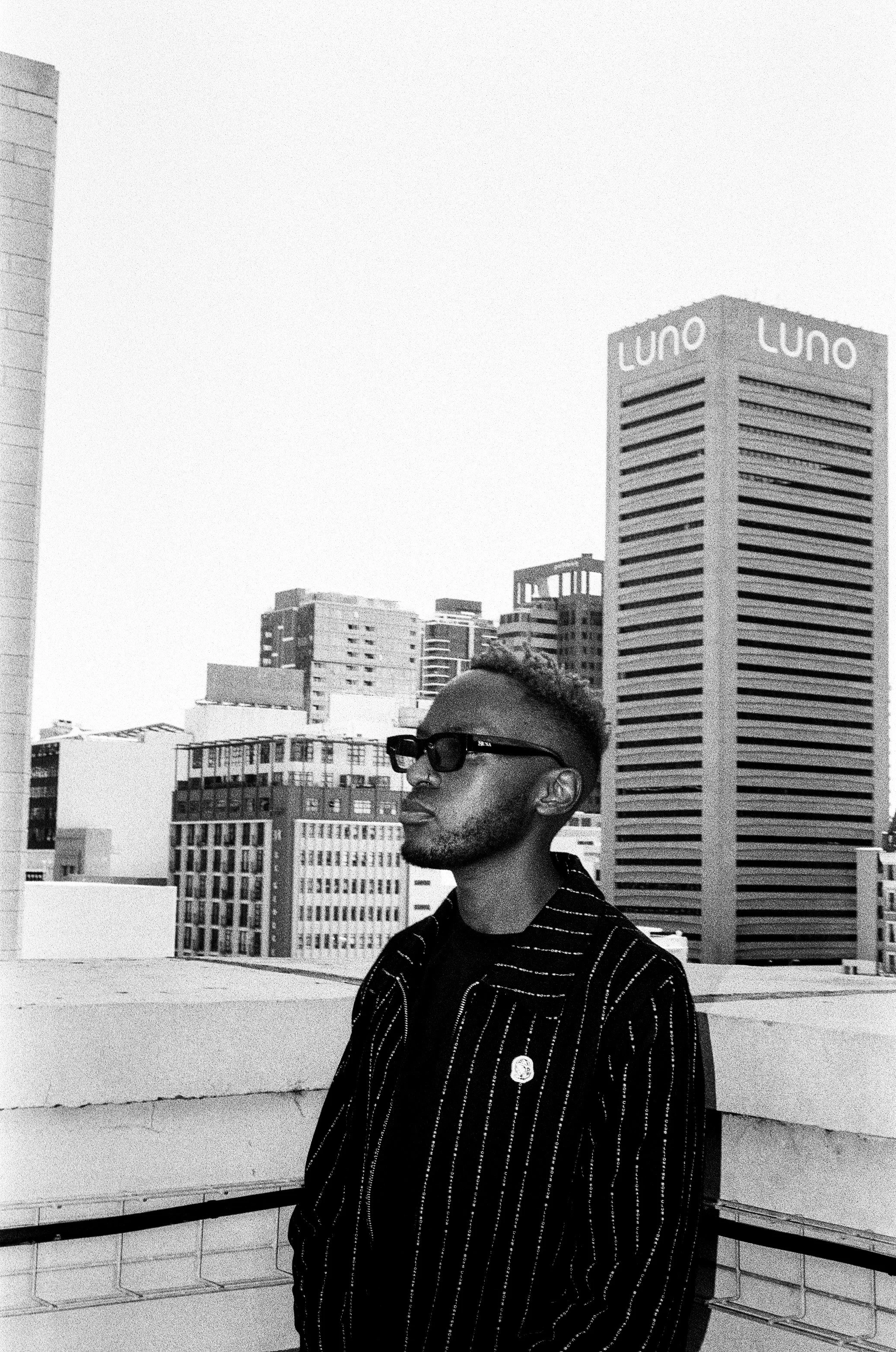 Themba Hadebe wearing sunglasses and a striped jacket stands on a city rooftop, with tall buildings and the "Luno" logo on a skyscraper visible in the background.