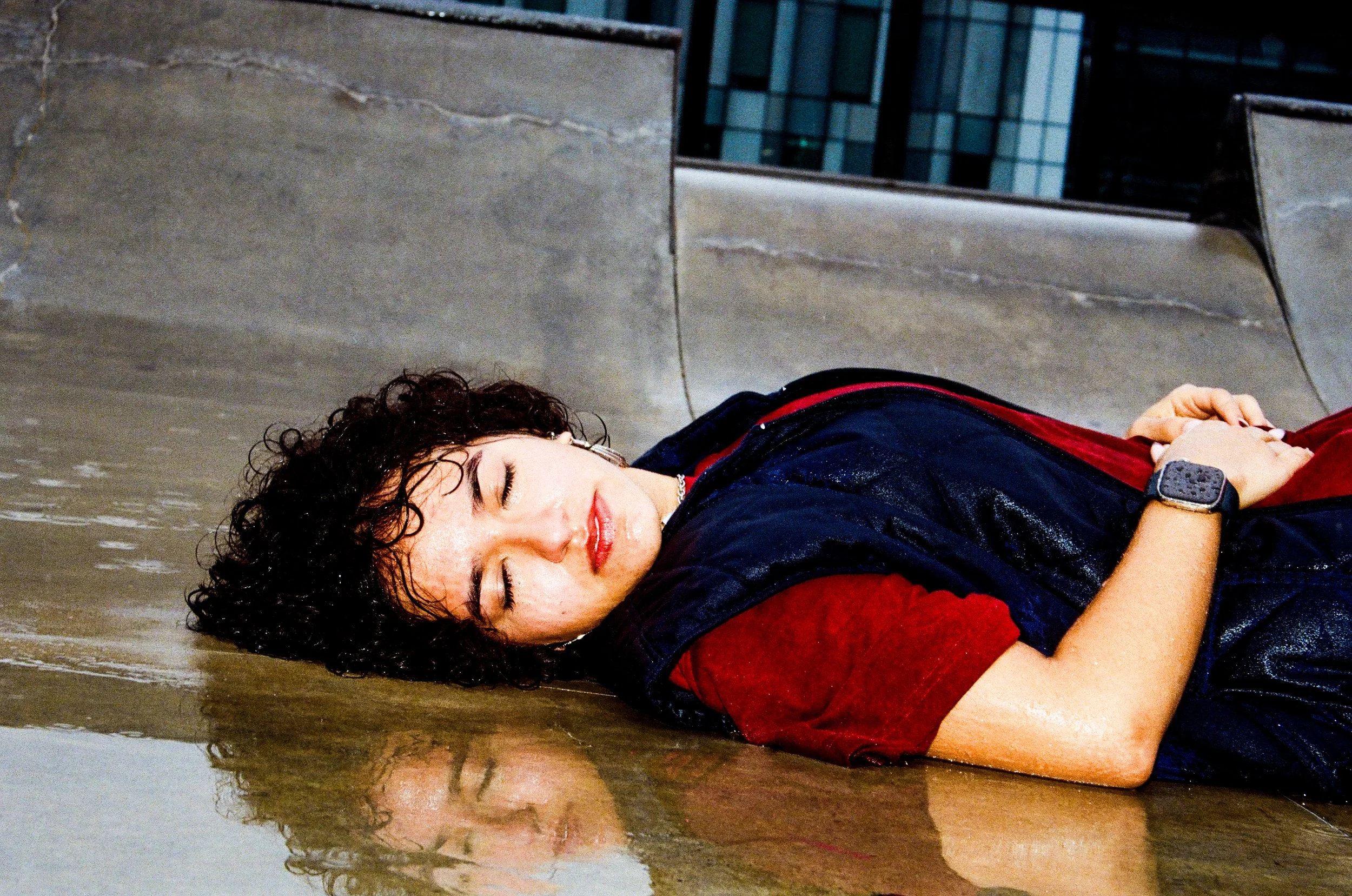 A woman with curly hair and a watch on her wrist lies unconscious on a wet surface, with her eyes closed and a peaceful expression.