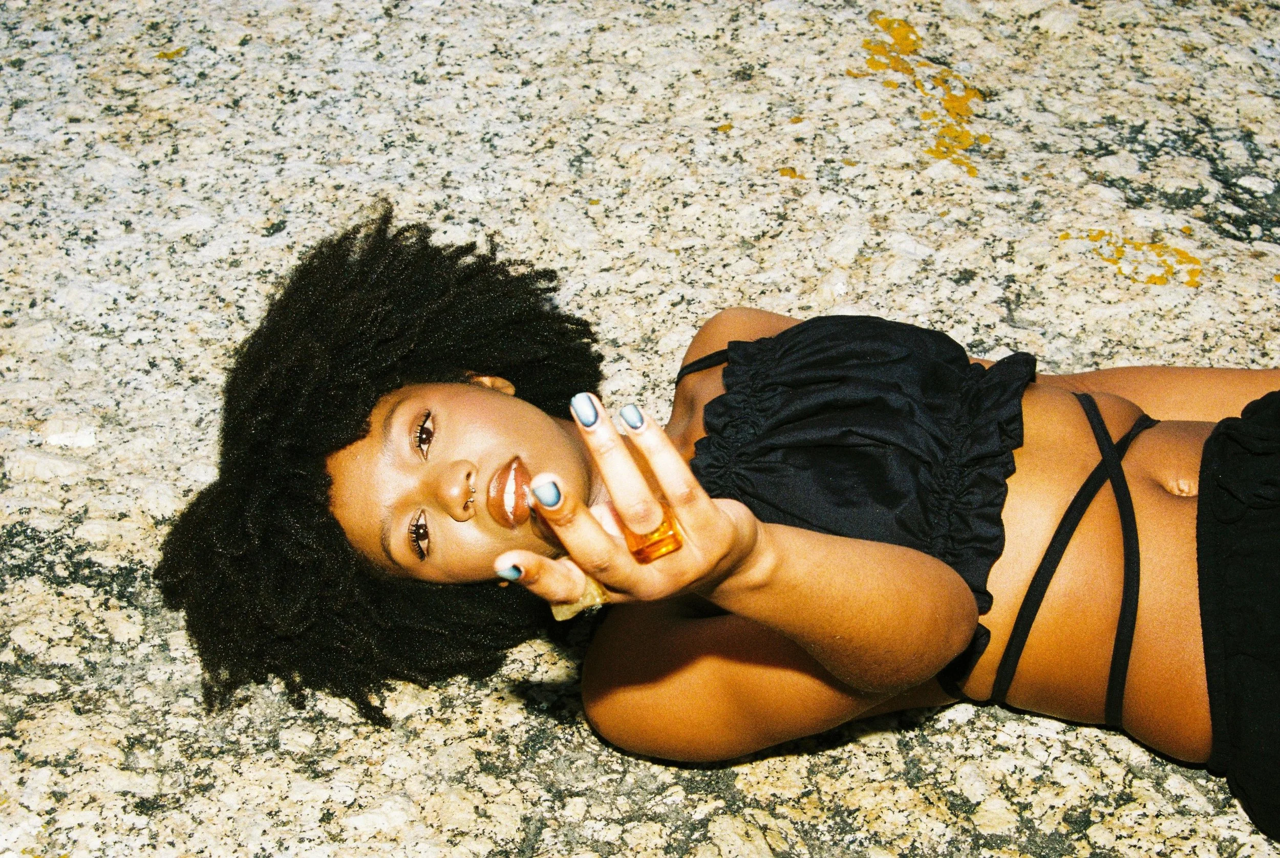 A young woman with curly black hair, lying on a granite floor, wearing a black crop top and shorts, holding up her hand with painted nails and a large amber ring, looking at the camera.