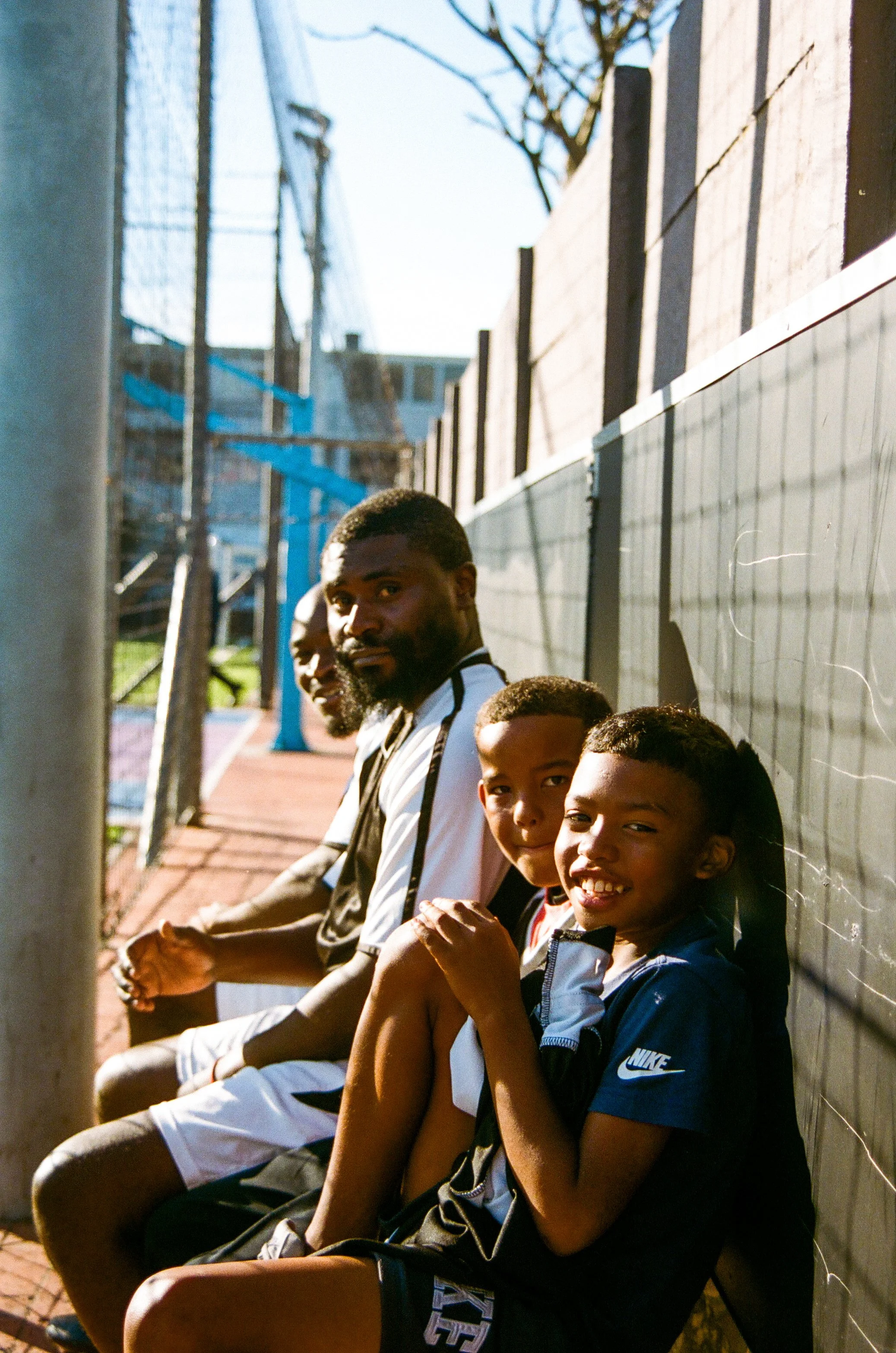 Four boys sitting on a bench beside a black fence, with sunlight illuminating their faces.