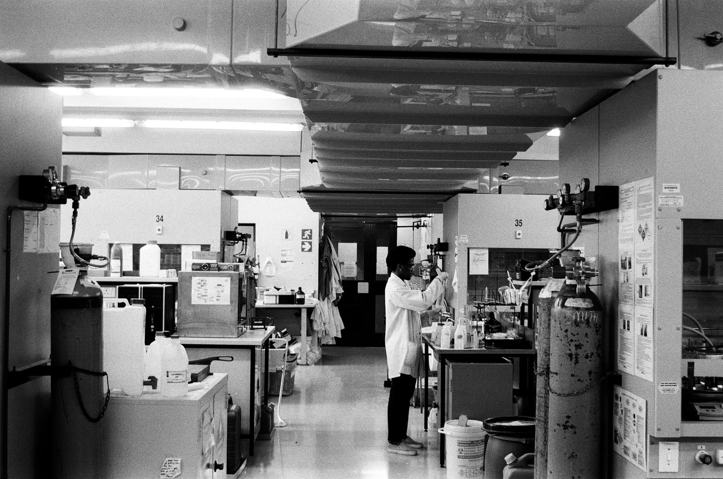 Black and white photo of a scientist working in a laboratory, surrounded by lab equipment, chemicals, and gas tanks.
