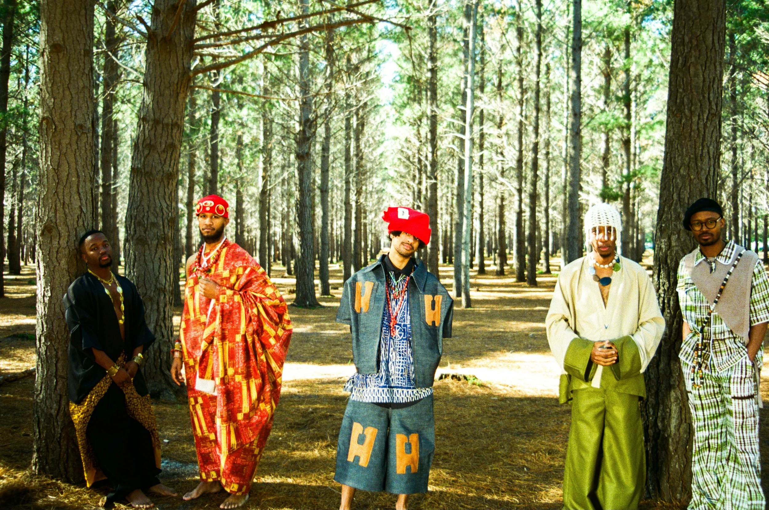 Kujenga standing in a forest with tall trees, dressed in a mix of traditional and modern clothing with colorful accessories.
