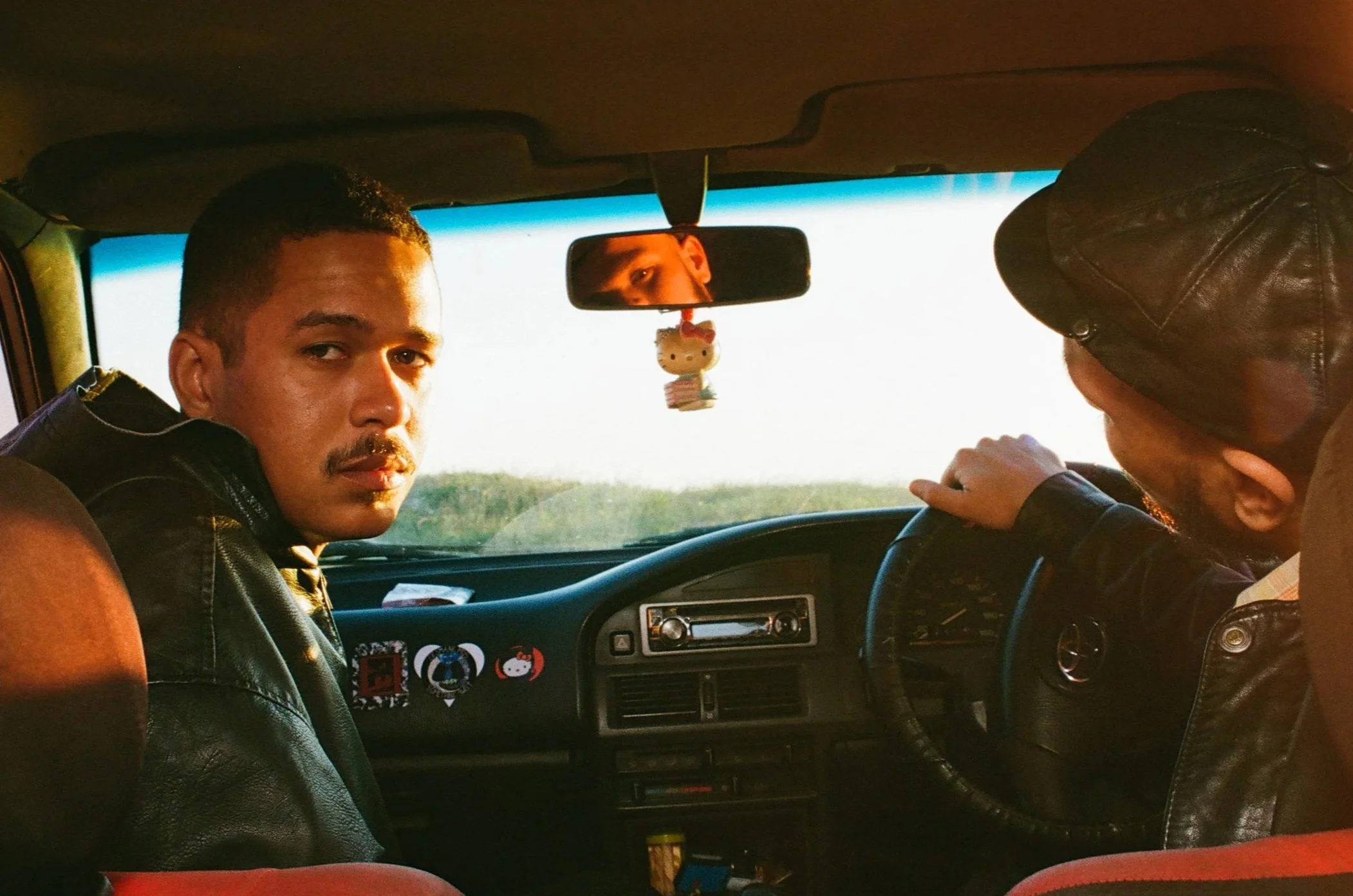 Two men sitting in the front seats of a car. On the left, Jarry Pitboi, is looking towards the camera, while the man on the right is driving. The car's dashboard and rearview mirror are visible, with a Hello Kitty air freshener hanging from it.