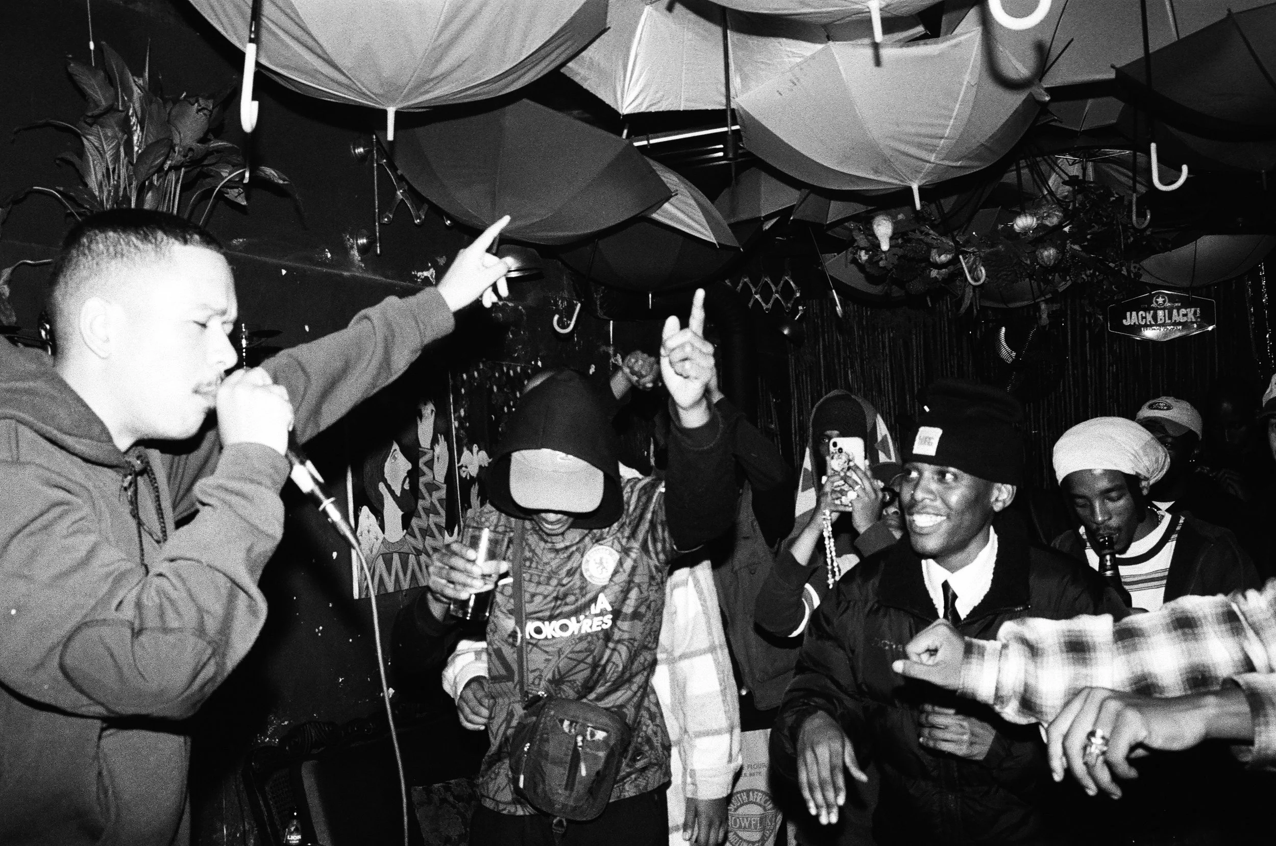 A black and white photo of Jarry Pitboi performing with a microphone in a club, surrounded by people dancing and taking pictures, with umbrellas hanging from the ceiling.