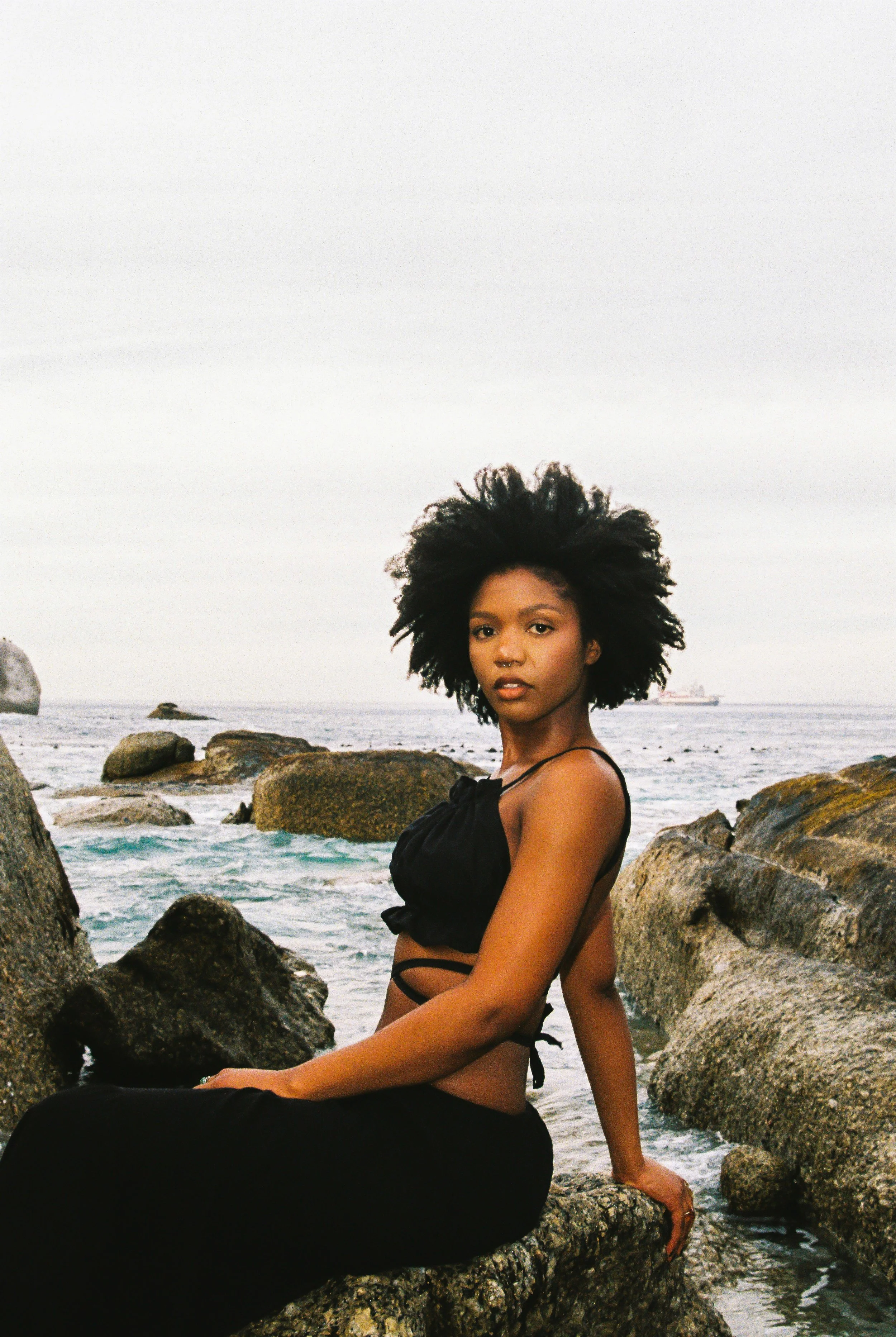 A young woman with natural afro hair sitting on rocks near the ocean, wearing a black outfit, with a ship visible in the distance.