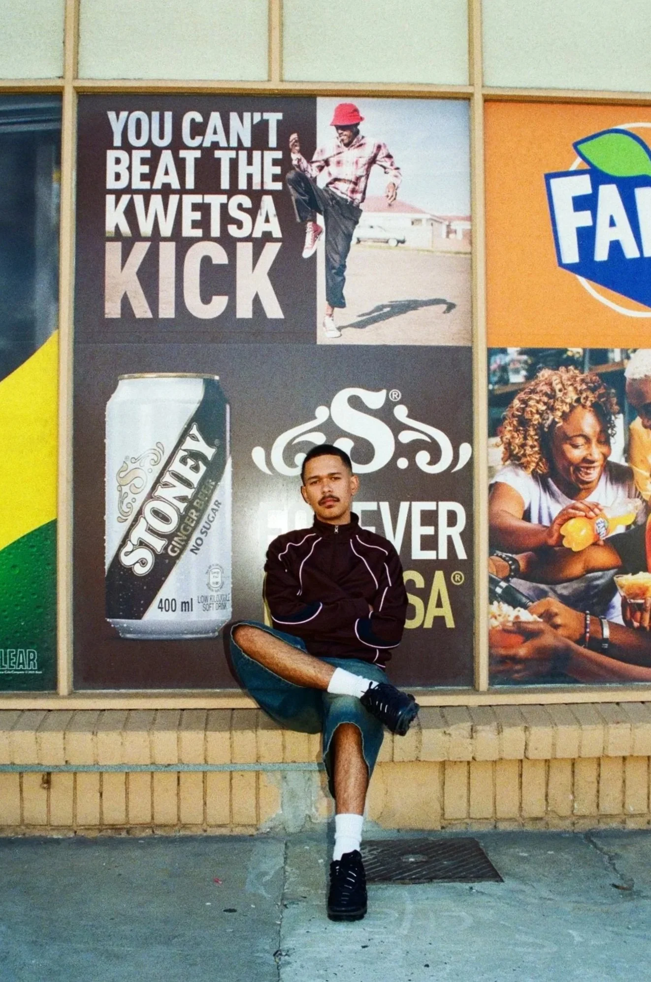 Jarry Pitbou sitting with crossed arms and one leg raised in front of a wall with advertisements, including a large poster of a can of Stone Ginger Beer, a sign with the words "You can't beat the Kwetsa kick," and a picture of a smiling woman eating.