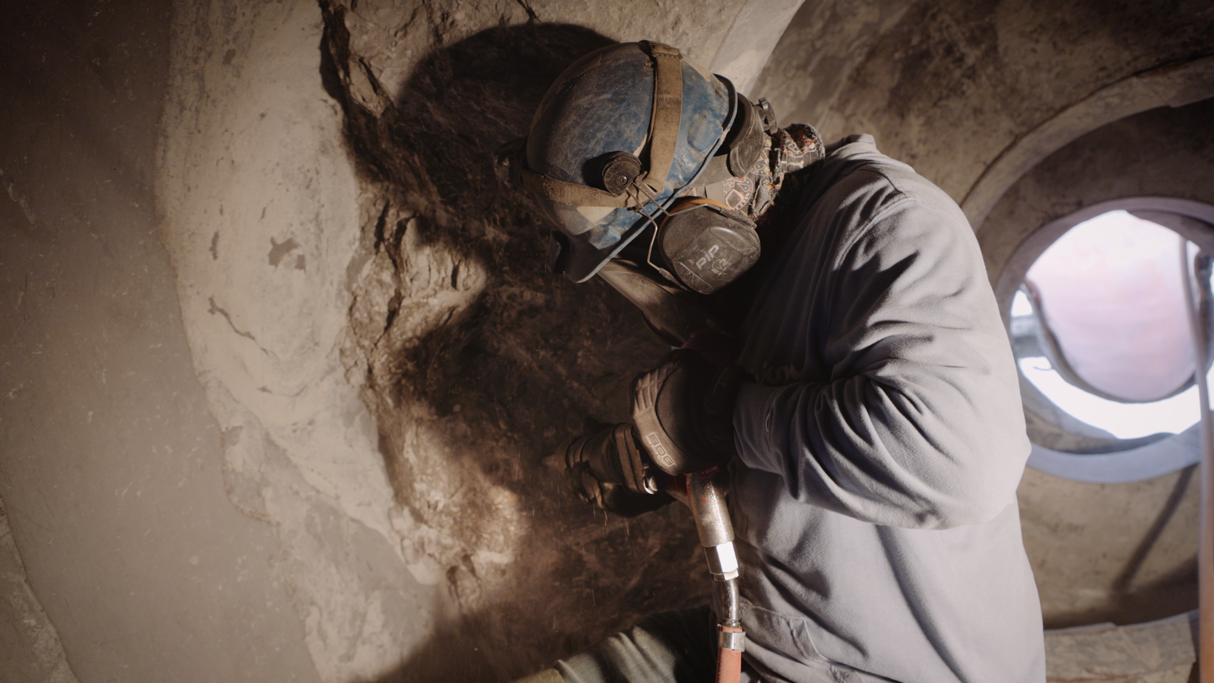 A worker in protective gear, including a hard hat, hearing protection, and gloves, using a jackhammer to remove concrete in order to repair a concrete truck drum.