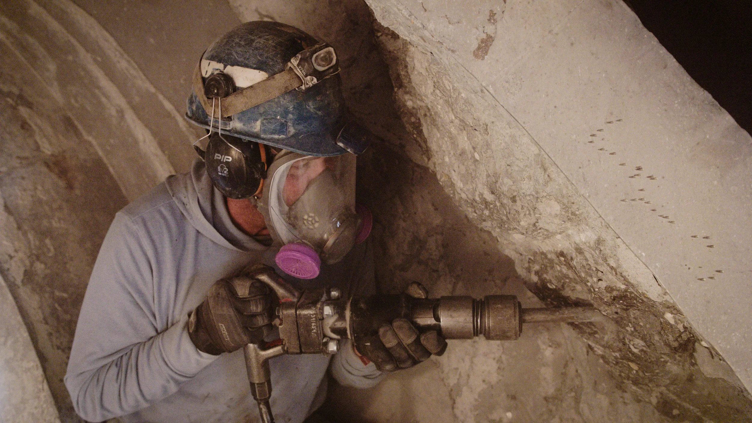 A worker wearing a blue hard hat, safety glasses, a respirator mask, and gloves using a jackhammer in a concrete drum.