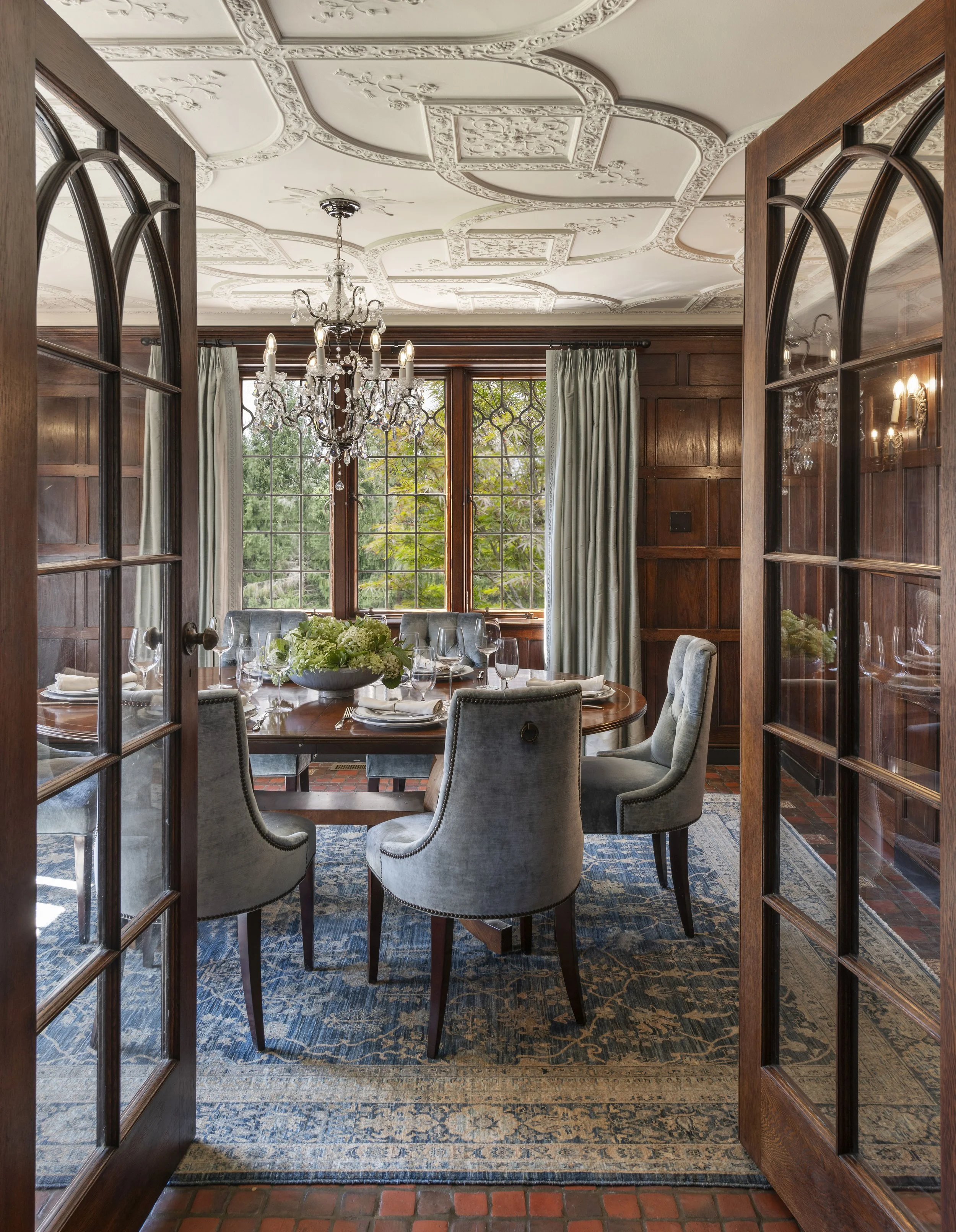 Tudor dining room with arched glass paneled oak doors, paneled walls, crystal chandelier, blue upholstered chairs, and custom oak dining table.