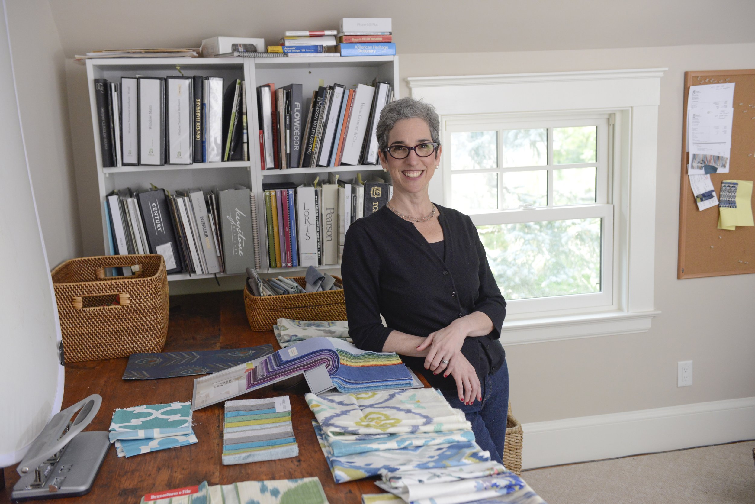 A woman with short gray hair, glasses, and a black shirt smiling indoors, standing next to a desk with fabric samples and color palettes, in front of a white bookshelf filled with books and binders, a window with greenery outside, and a bulletin board on the wall.