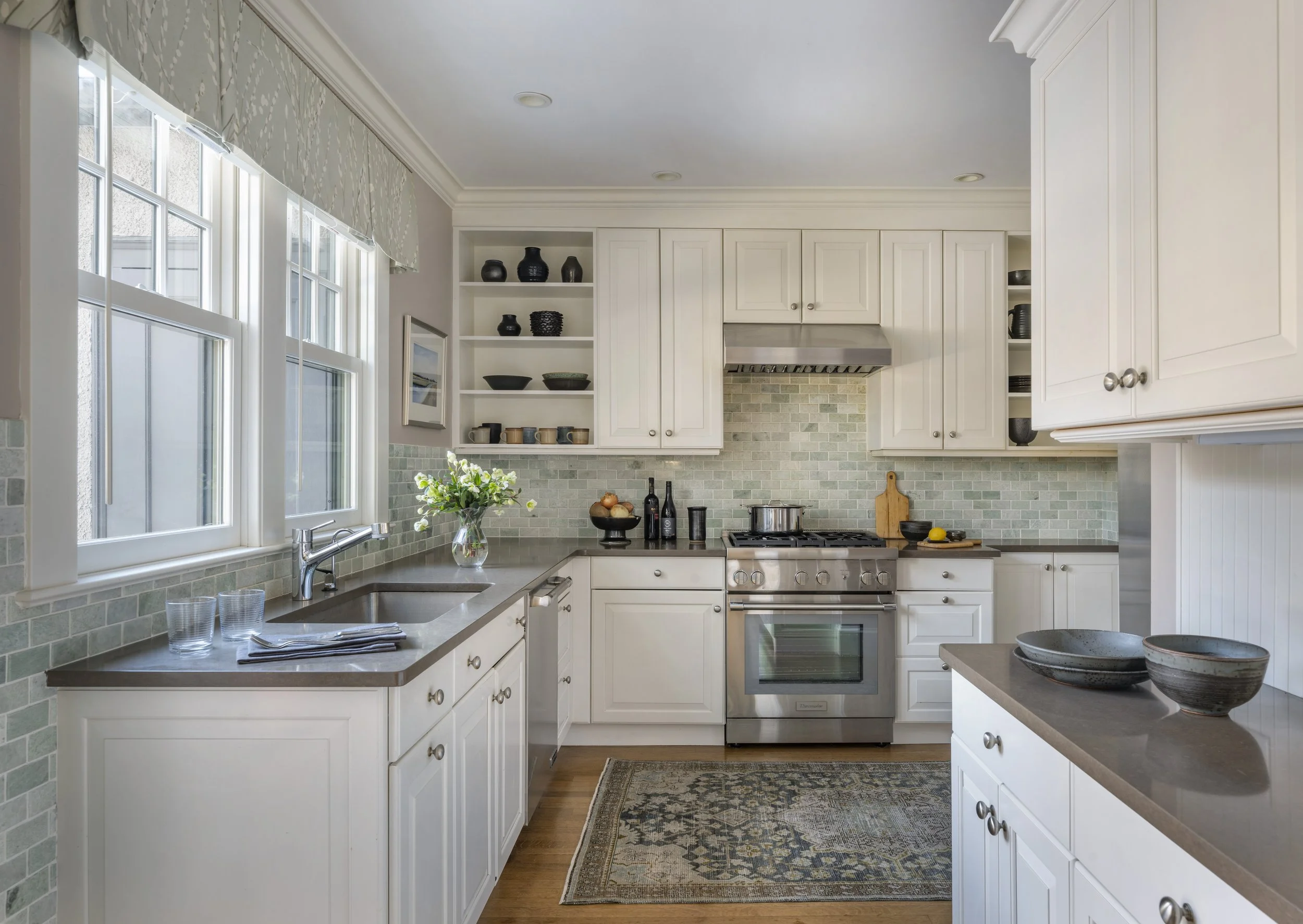 Cambridge kitchen designed by AbbeyK with white cabinetry, green marble tile backsplash, open shelving with ceramic dishware, a valance over the sink, Persian rug, and stainless range.