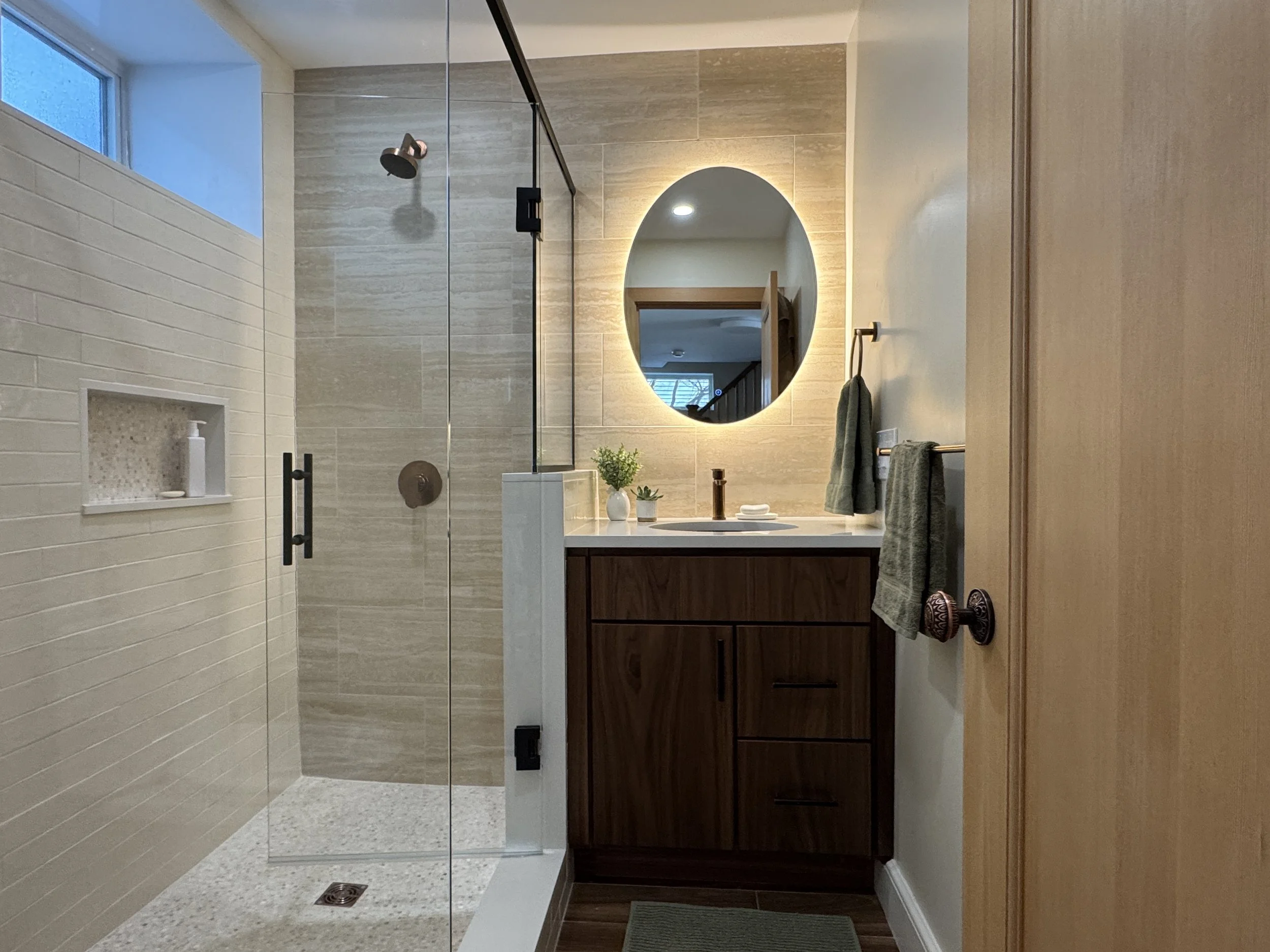 Custom basement bathroom designed by AbbeyK Interior Design in Lexington MA featuring walnut vanity, backlit oval mirror, travertine tile, glass shower enclosure with pebble floor, and copper fixtures.