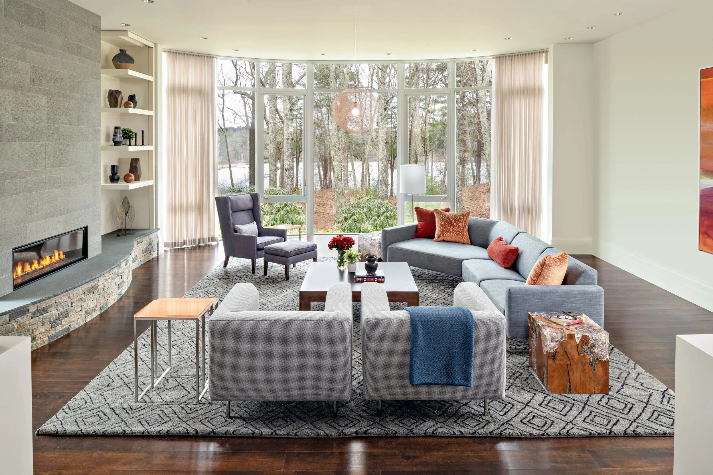 Modern, Bright living room with large windows showing trees outside. Contains a gray sectional sofa with red and orange pillows, a gray armchair, two smaller gray chairs with a blanket, a wooden coffee table, and a fireplace with a stone base. Decor