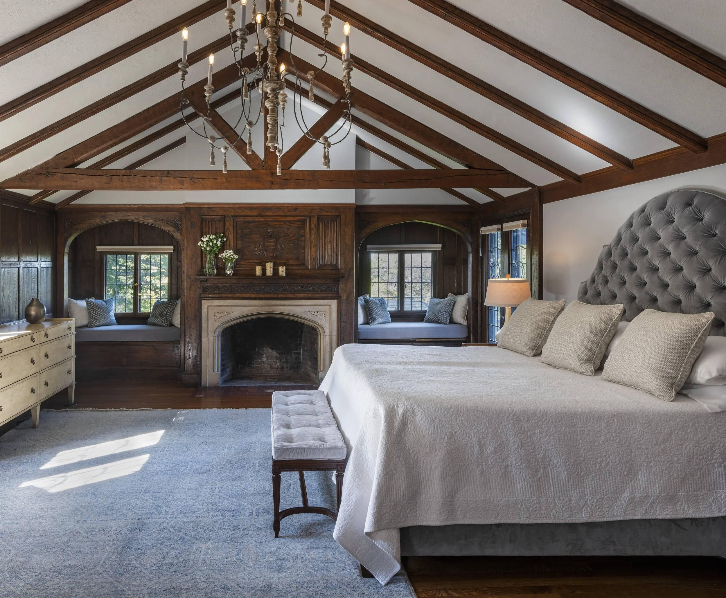 A spacious bedroom with a vaulted ceiling and exposed wooden beams. Features include a large bed with tufted gray headboard, white bedding, and multiple pillows. There is a bench at the foot of the bed, a lamp on the side table, and built-in window seats with cushions and pillows on either side of a stone fireplace with wood paneling and windows with decorative framing.