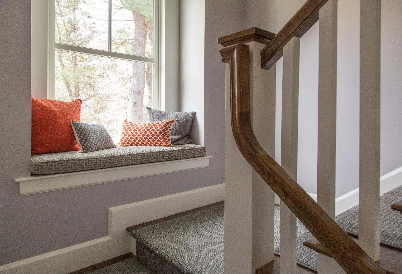 Needham staircase with walnut-stained railing, dark stair runner, and window seat with orange geometric pillows.