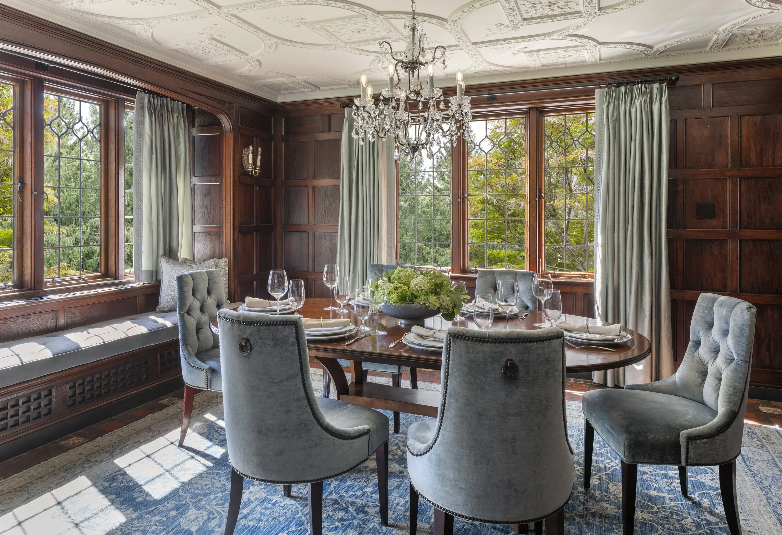 Elegant dining room with wooden paneled walls, large windows with green curtains, a chandelier, a round wooden table set with white napkins, plates, wine glasses, and a floral centerpiece, and upholstered chairs.