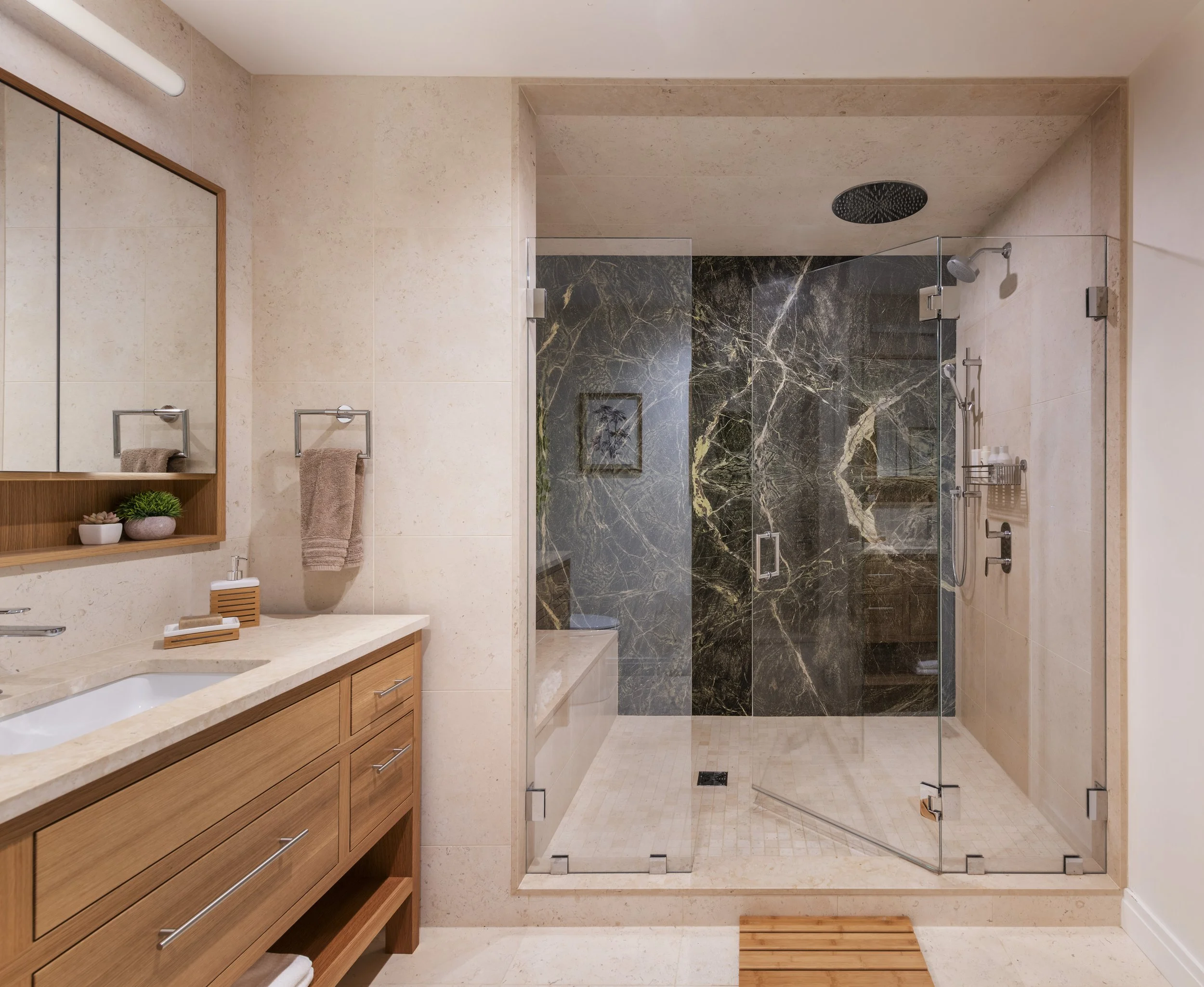 Seaport primary bathroom with custom wood vanity, integrated mirror storage, linear lighting, slab stone shower wall, and frameless glass shower enclosure.