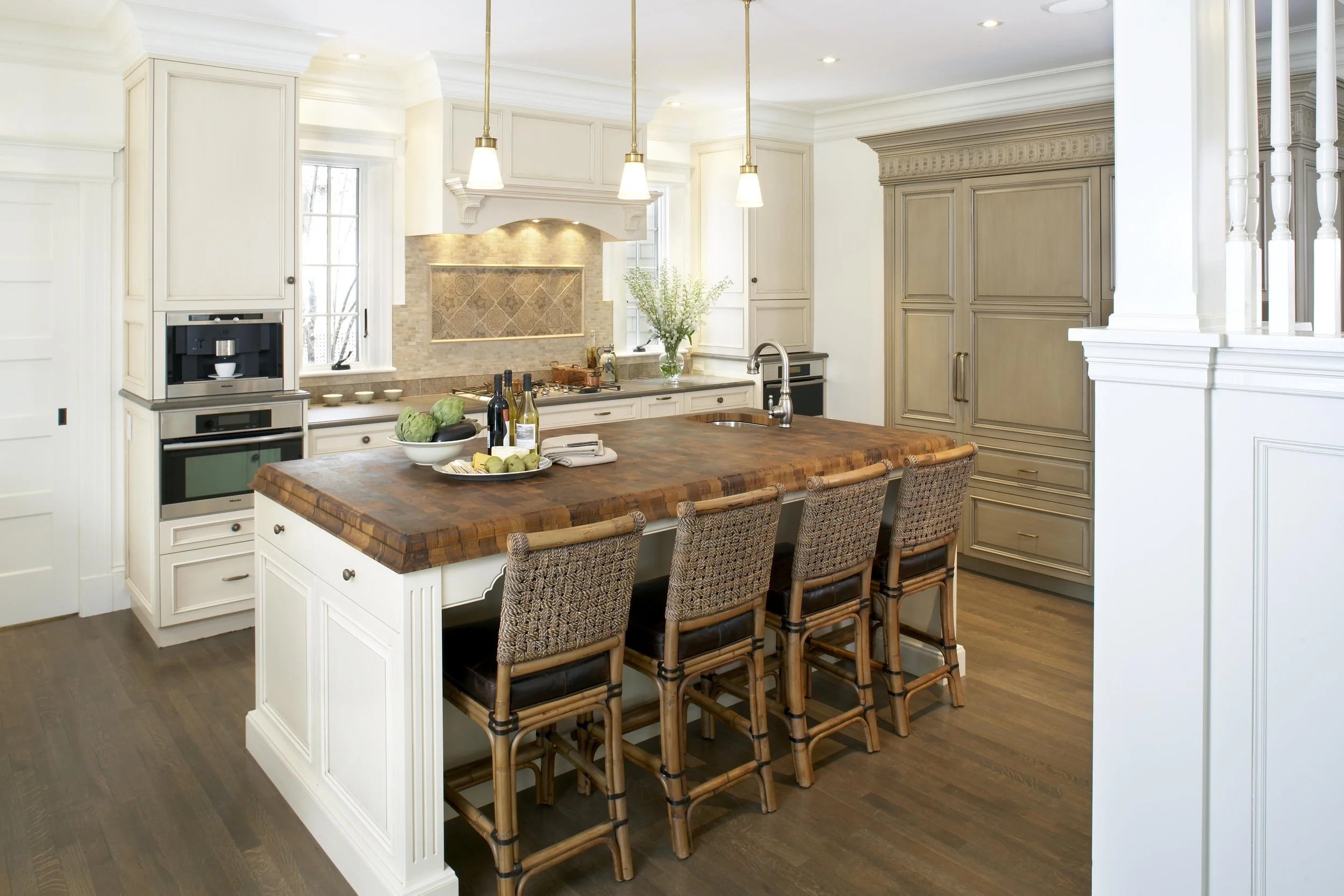A modern farmhouse kitchen with a large island, wooden countertop, beige cabinetry, and stainless steel appliances. Four woven chairs are at the island, and the kitchen is brightly lit with pendant lights.