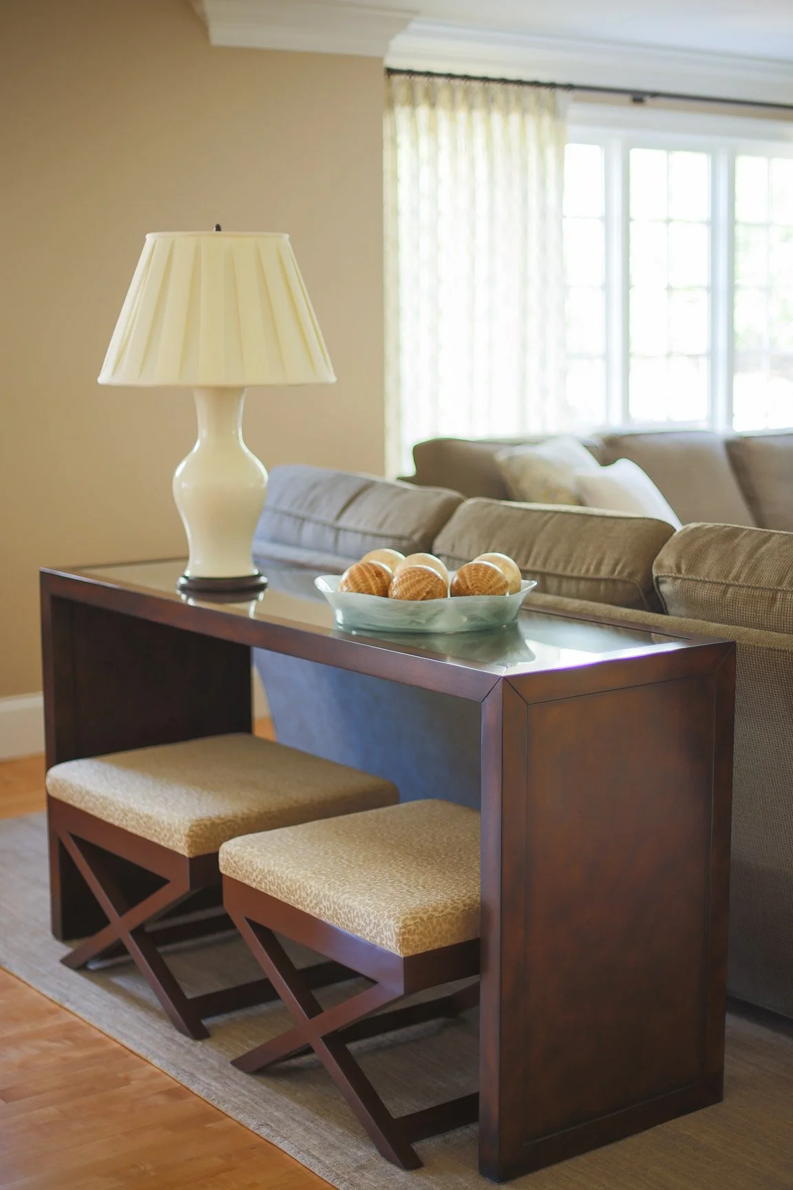Living room with a side table holding a lamp and a bowl of shells, beige sofa, large windows with curtains.