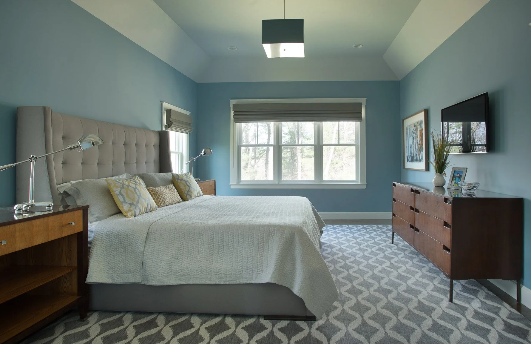 Needham primary bedroom with tall upholstered headboard, custom wood dresser, squared ceiling light, bold patterned rug, and patterned pillows.