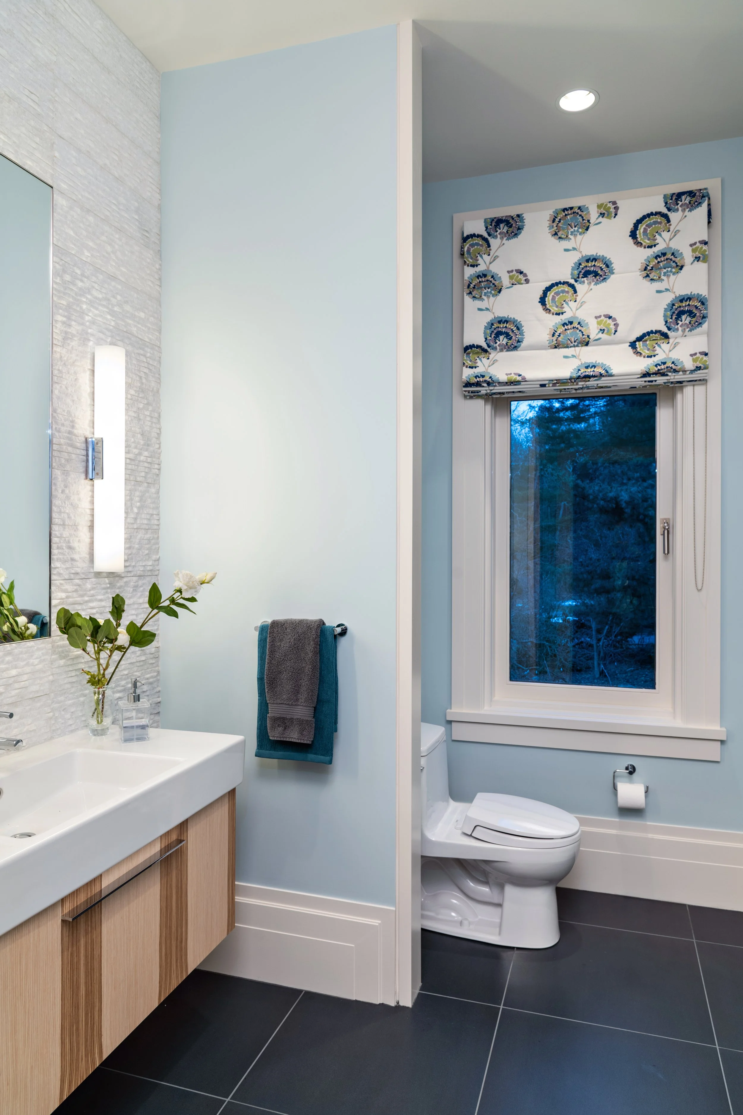Lincoln modern bathroom with soft blue walls, floating wood-grain vanity, white rectangular sink, textured stone tile, chrome vertical sconces, floral Roman shade, and charcoal floor tile.