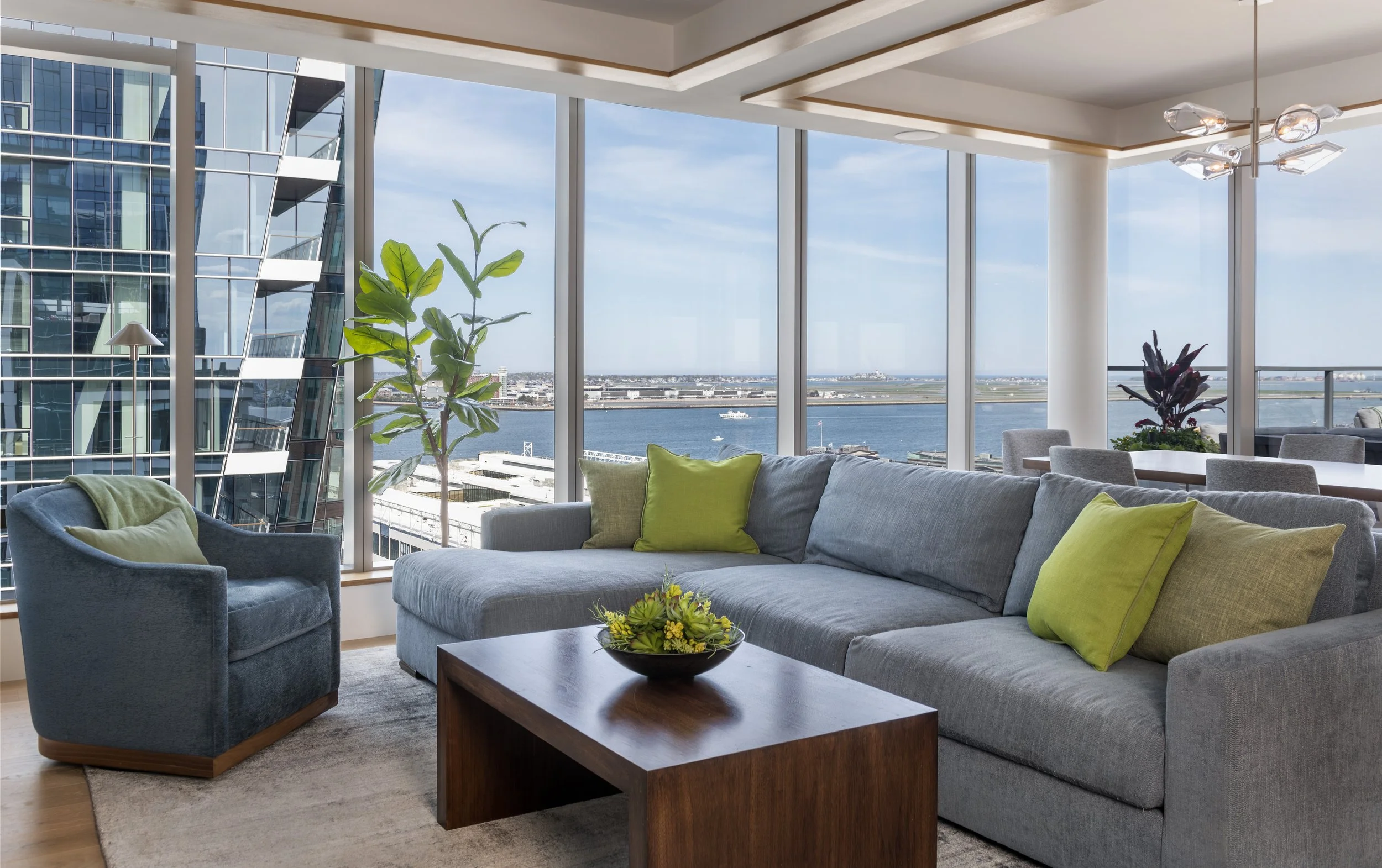 Modern living room with large windows overlooking the boston harbor  modern blue sofa with green pillows, blue armchair with green pillow, walnut coffee table with a floral centerpiece, indoor plants, contemporary lighting fixture, and a dining area.