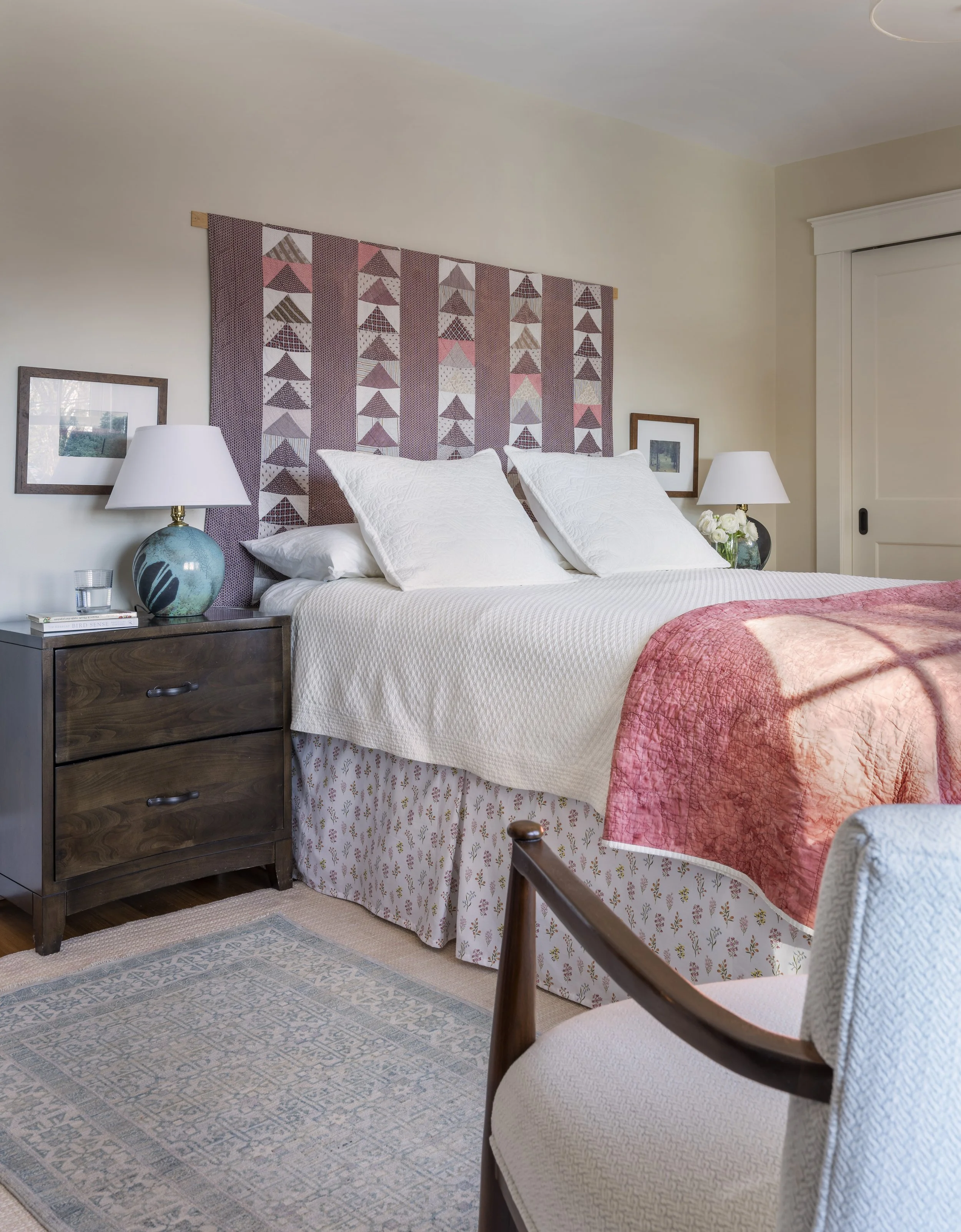 Cambridge bedroom designed by AbbeyK with an antique quilt used as a headboard, a pink quilt layered on the bed, a vintage wood nightstand, and an artisanal raku ceramic lamp.