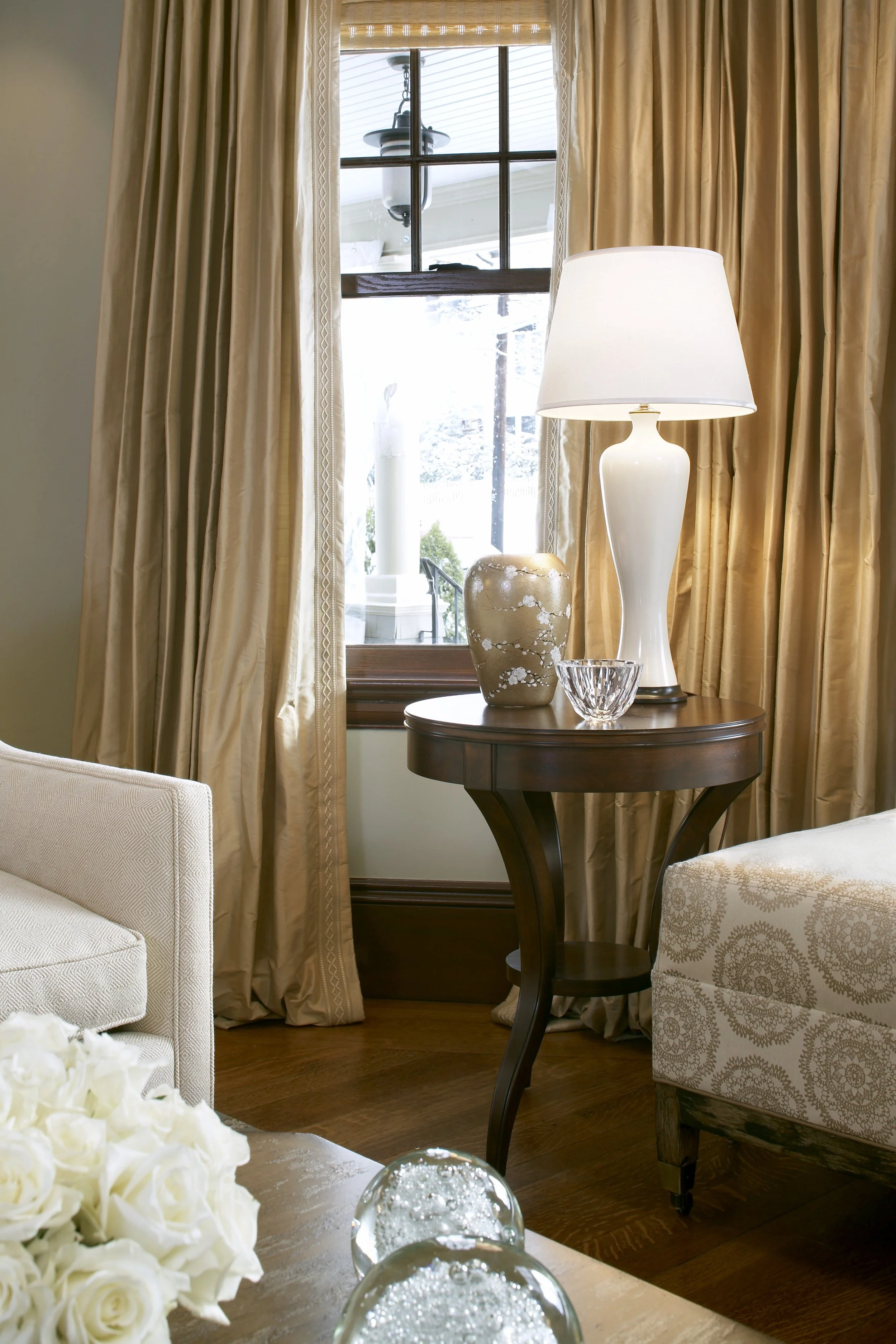 Living room detail with white lamp, gold vase, and silk drapes on a round wood side table featured on This Old House.