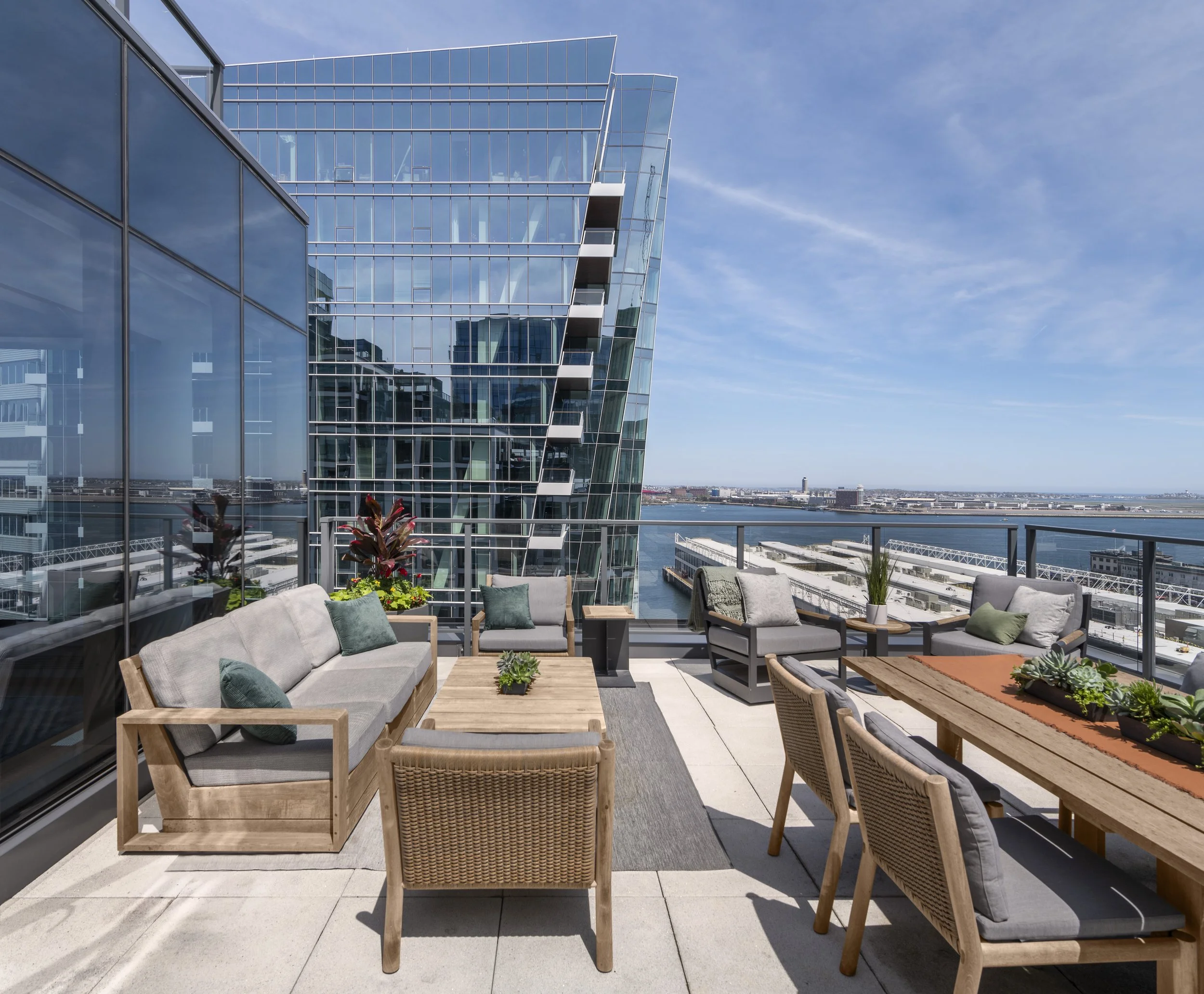 Seaport roof deck with teak seating area, dining area, woven lounge chairs, lush planters, and panoramic harbor views.