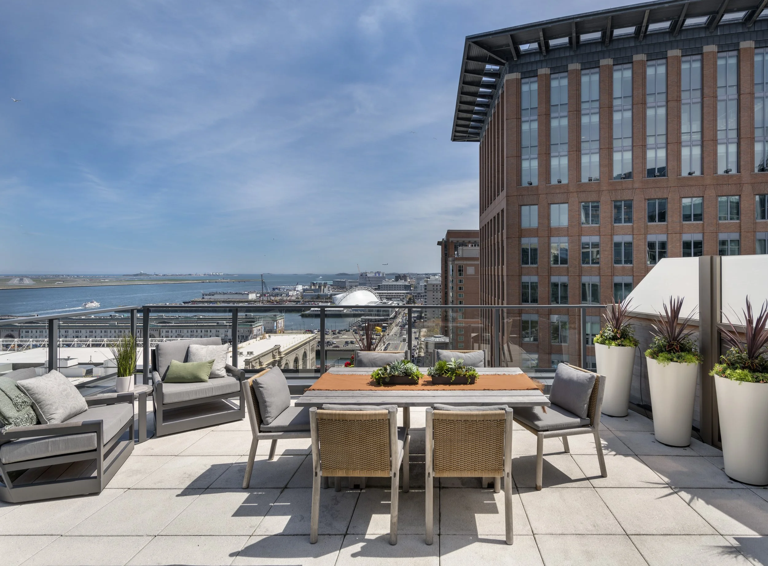 Roof deck with teak sofa, woven chairs, dining area, layered planters, and skyline and harbor views.