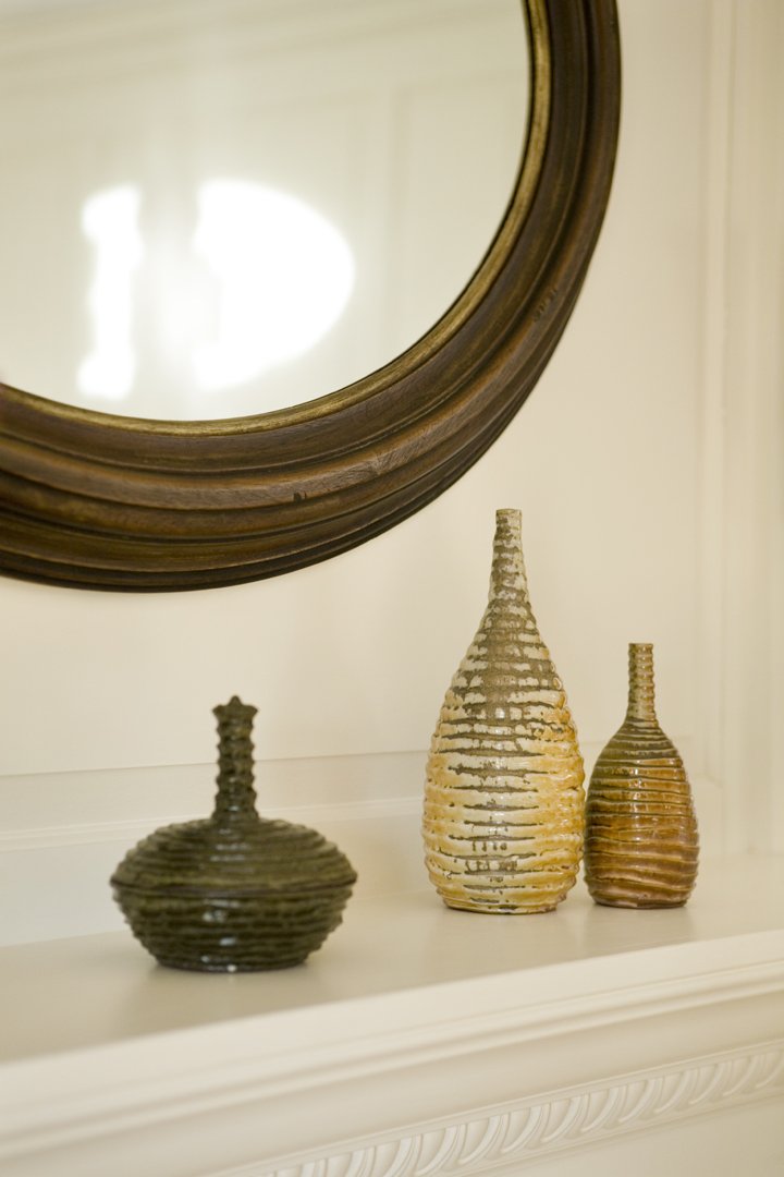 Three decorative vases on a mantle in Lexington on a mantle place with textured, horizontal ridges and narrow necks on a white mantle in front of a large, round, wooden-framed mirror.