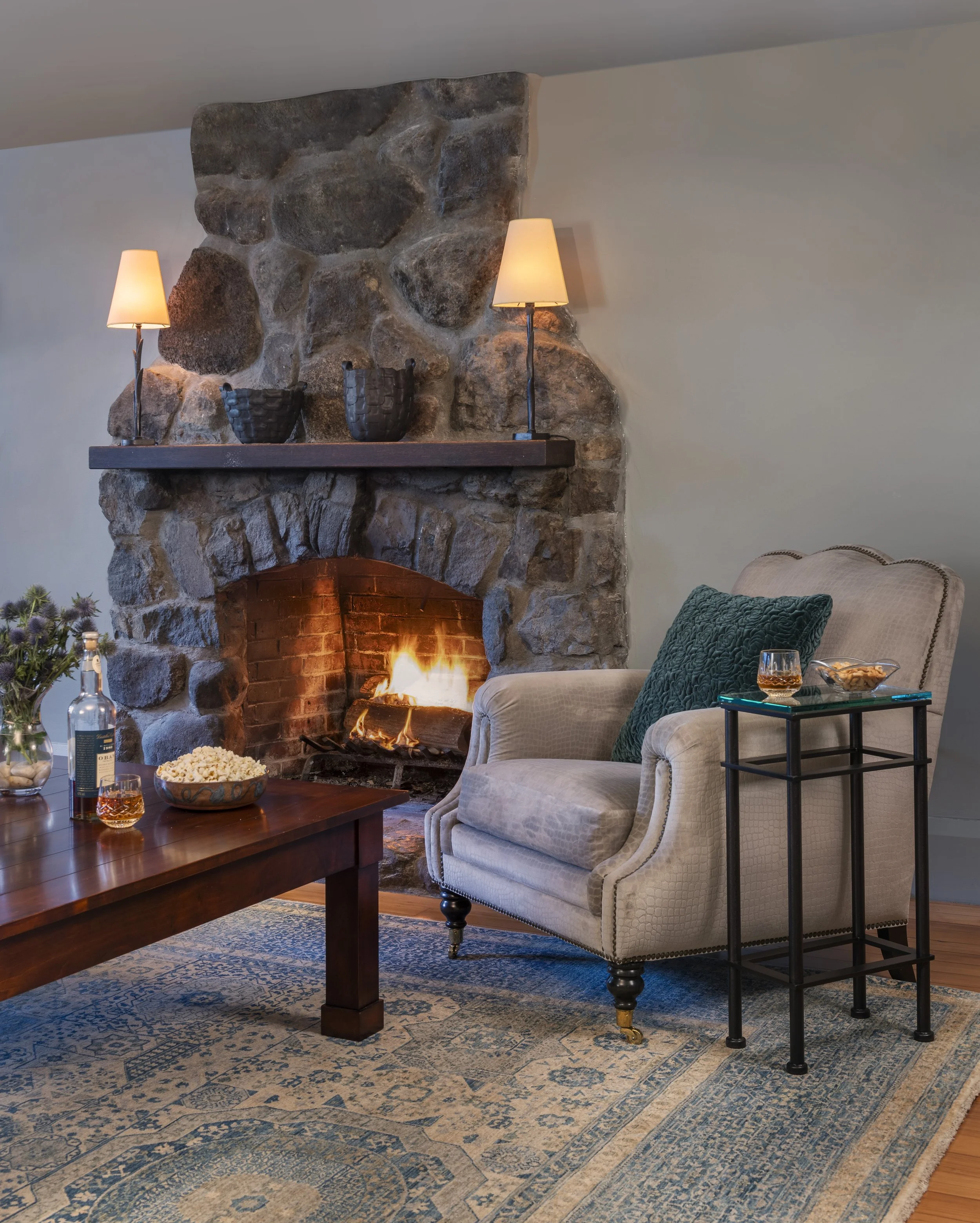 Tudor stair hall with carved oak staircase, terracotta tile floor, modern runner rug, and view to dining room.
Lower-level Tudor family room with stone fireplace, walnut square coffee table, taupe armchair, teal accents, Mamluk rug, and oak floors.
