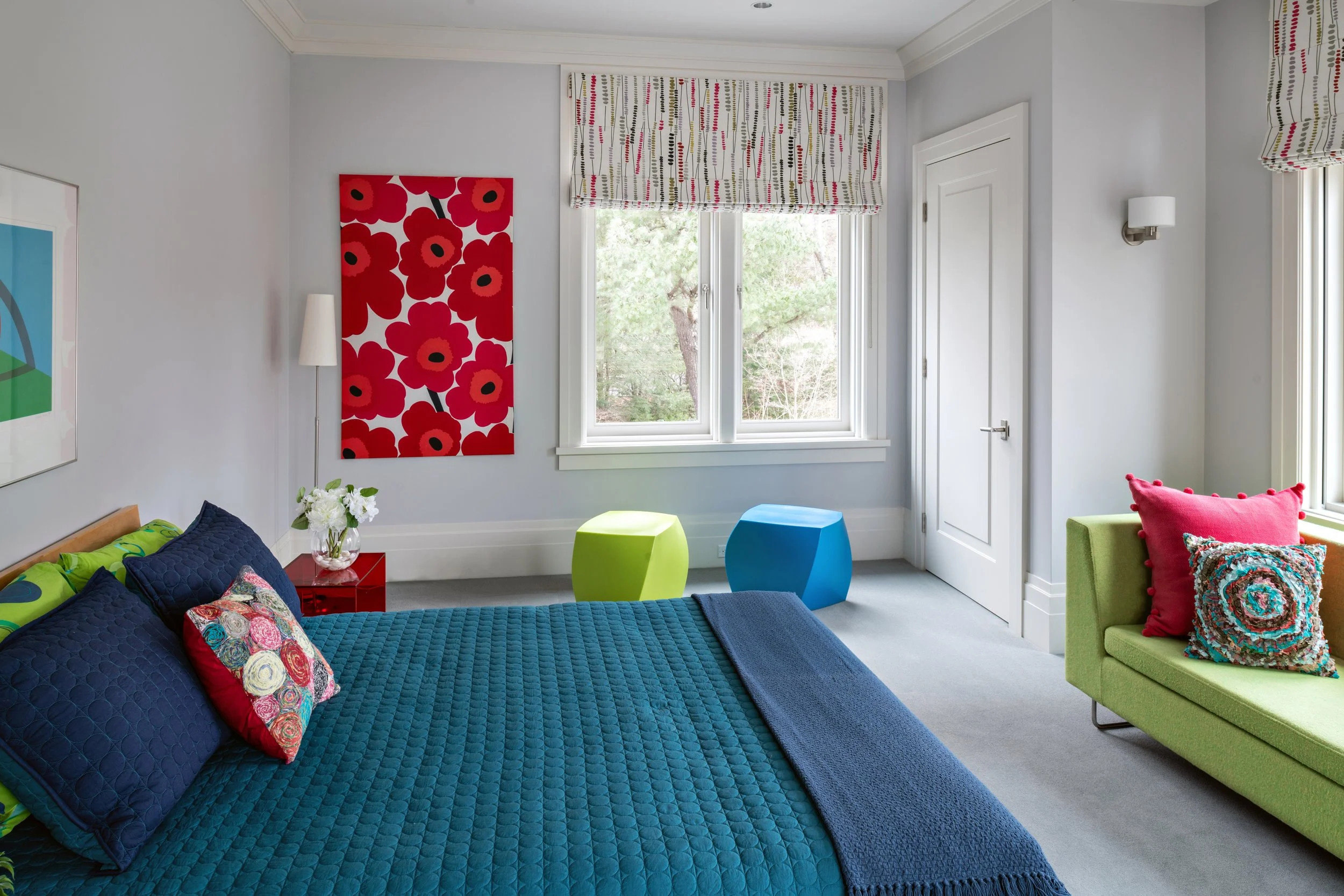 Lincoln guest bedroom with teal and navy bedding, Marimekko red floral art, colorful trimmed Roman shade, chartreuse and blue cube stools, green lounge chair, gray walls, and wool wall-to-wall carpet.
