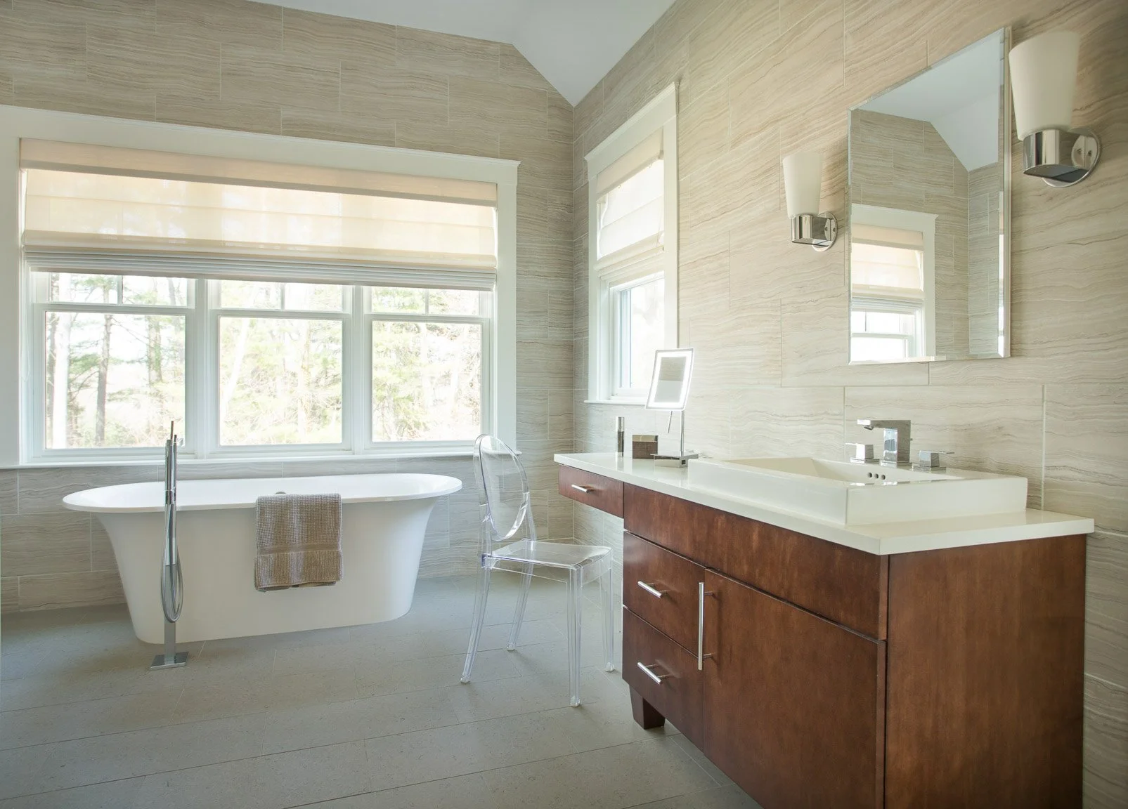 Needham primary bathroom with freestanding tub under large window, walnut vanity, ivory porcelain floors and walls, glass shower enclosure, mosaic tile wall, and a clear Ghost chair.