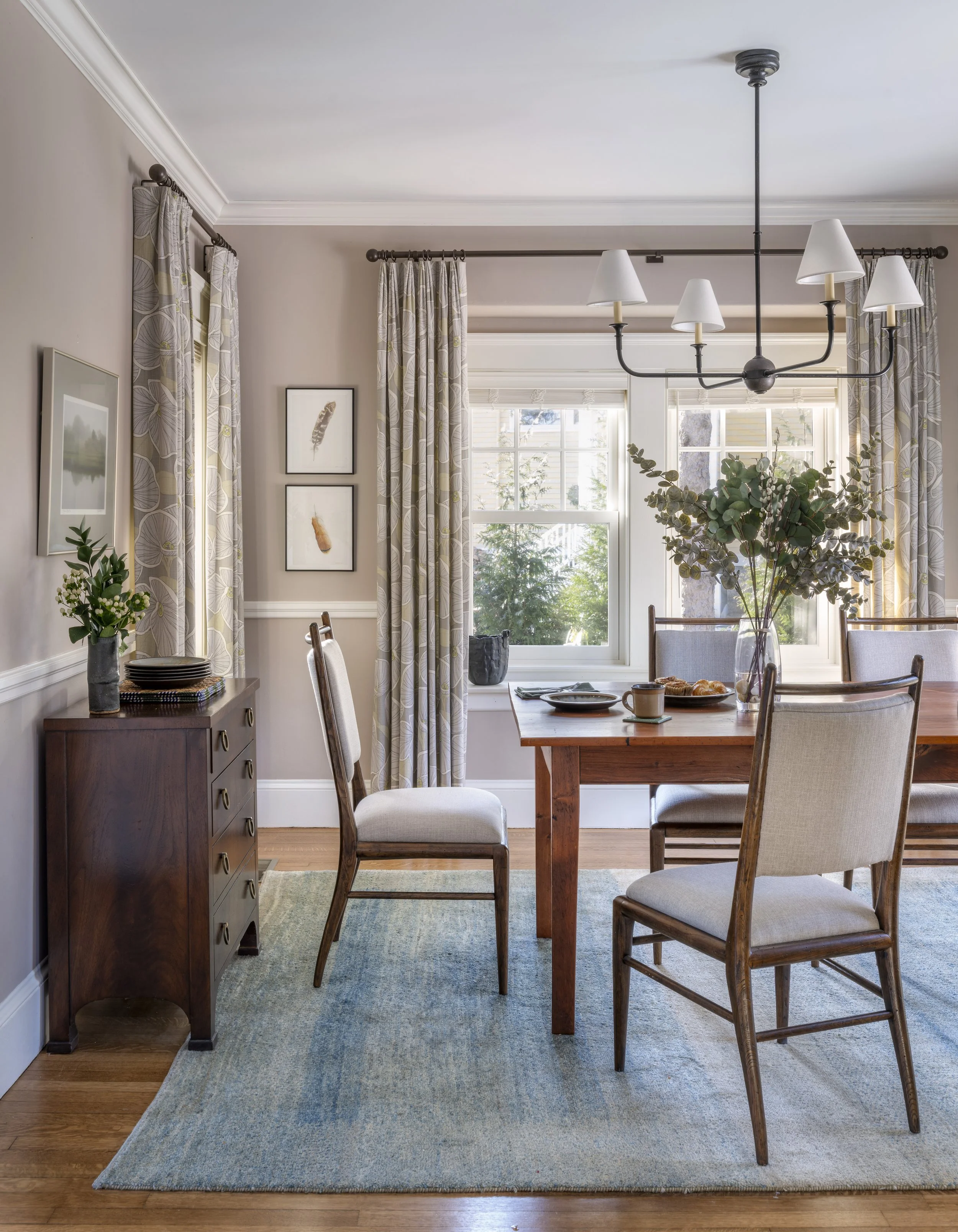 Vertical view of the Cambridge dining room designed by AbbeyK showing custom window panels with wrought iron hardware, a chandelier with fabric shades, vintage dining chairs, and framed artwork.
