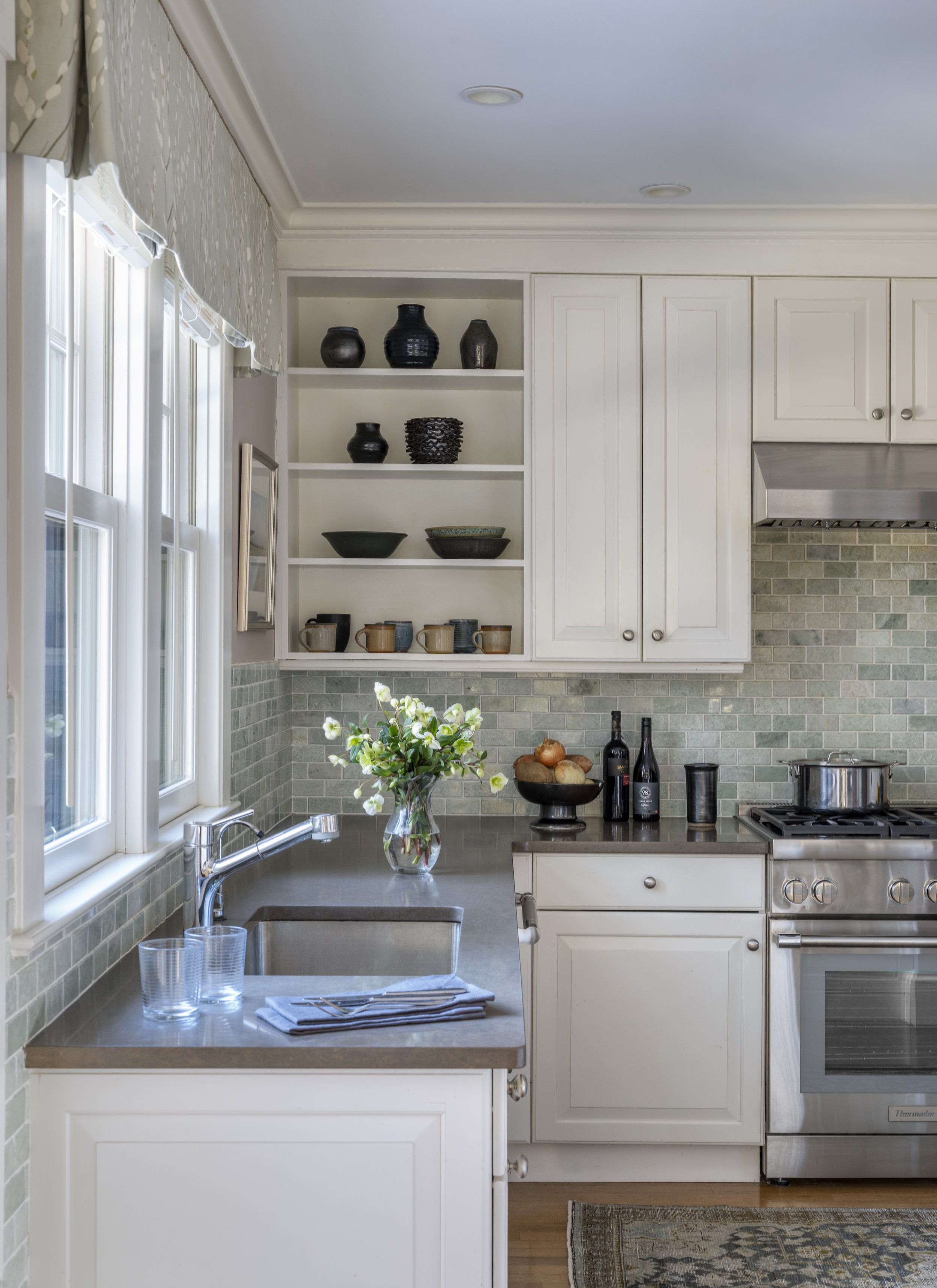 Kitchen detail in the Cambridge home showing open shelving with ceramic dishware, green marble tile backsplash, and a tailored valance over the sink, designed by AbbeyK.