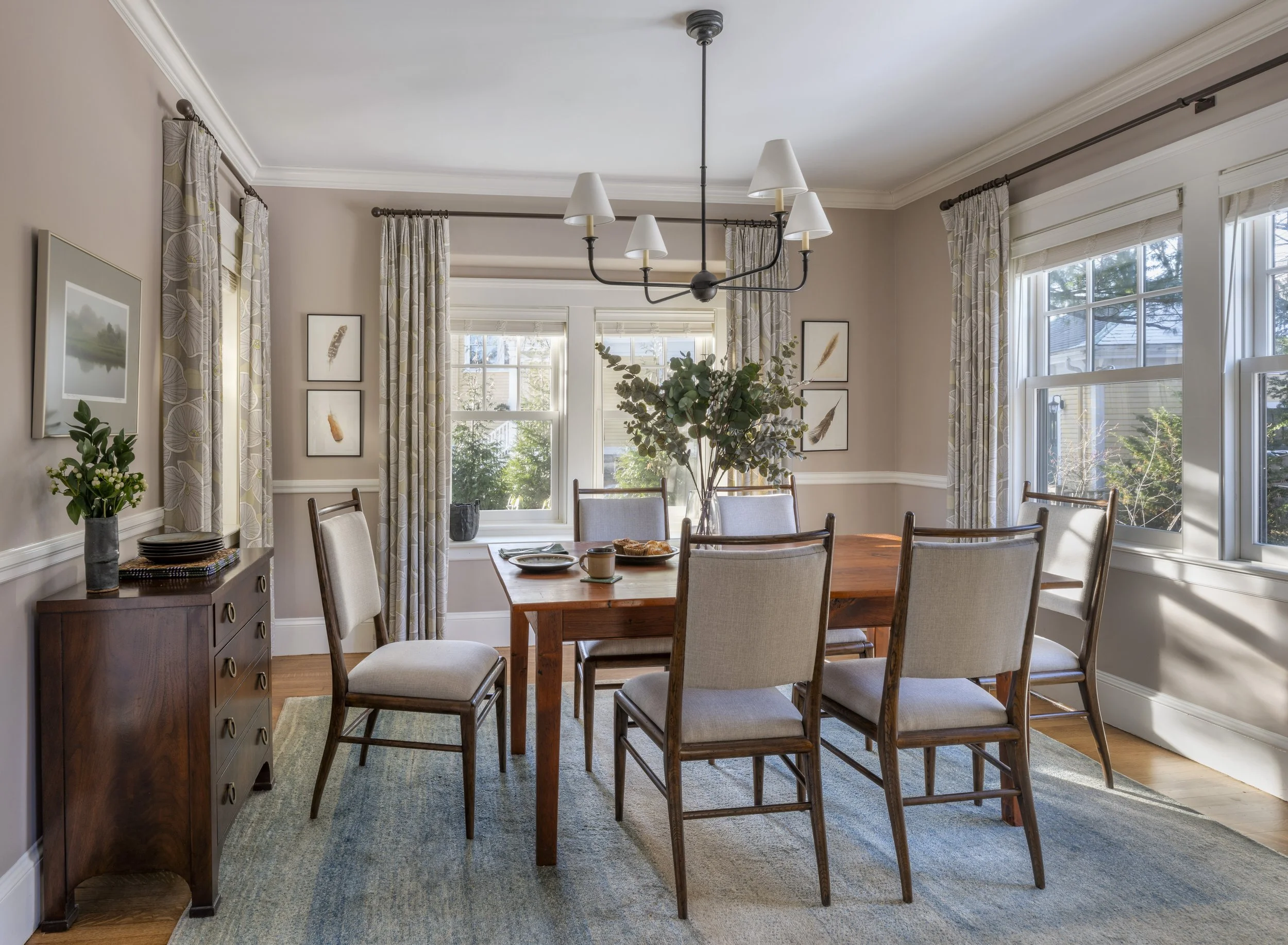 Dining room with reclaimed table, wood framed dining room chairs with ivory upholstery, a small vintage server, custom large scale gray drapes with leaves, art with feathers, wrought iron chandelier with shades bringing the outside in