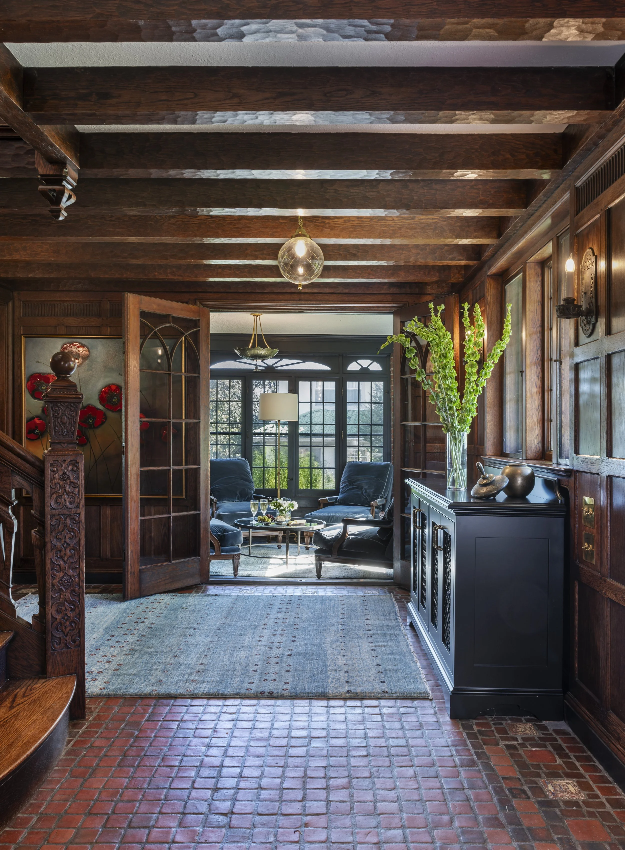 Tudor entry hall with exposed oak beams, terracotta tile floor, modern runner rug, carved wood paneling, and view into sitting room.