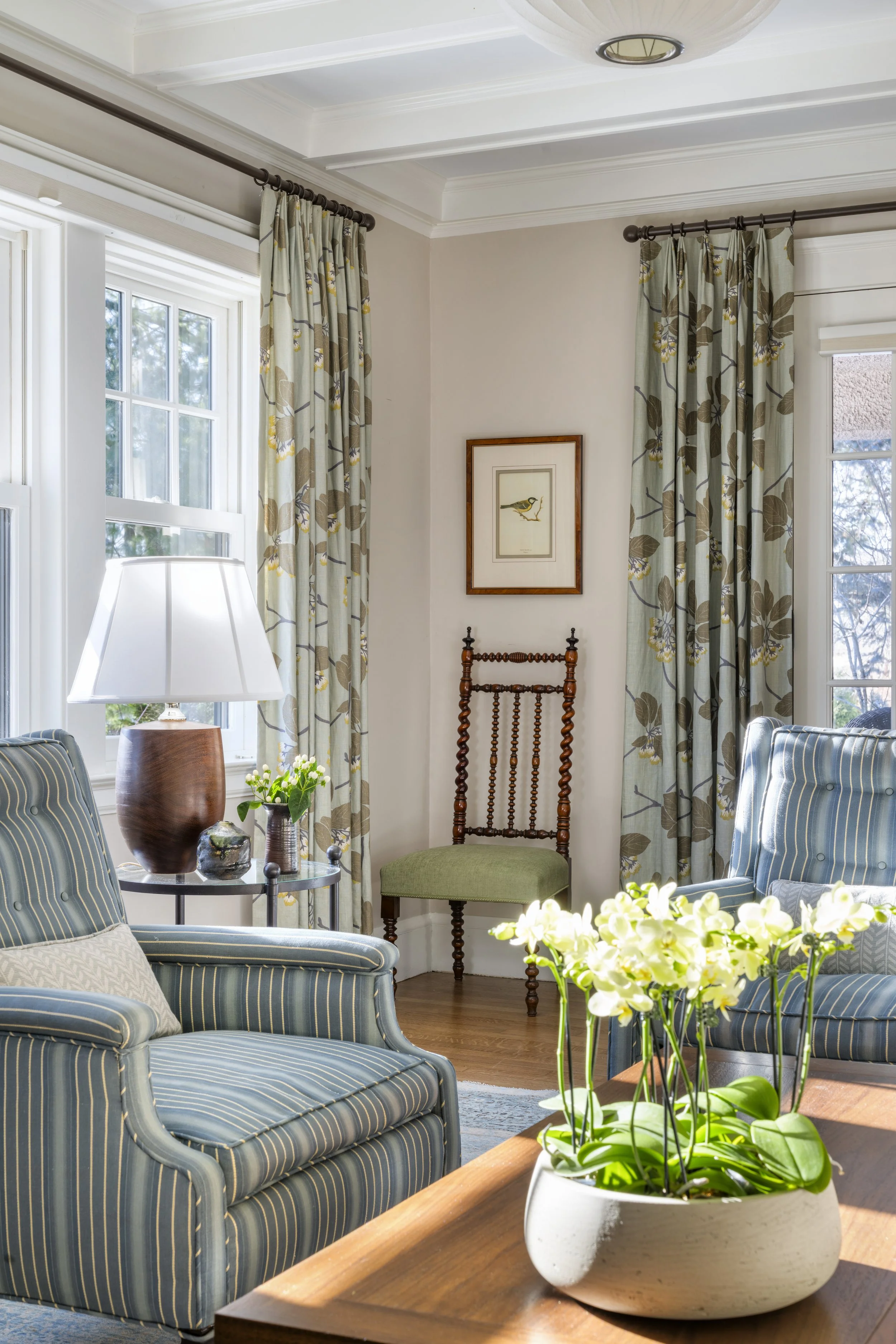Living room detail in the Cambridge home showing blue striped vintage chairs, a custom walnut Ming-style coffee table, a wood lamp on a glass side table, and large-scale patterned window treatments, designed by AbbeyK.
