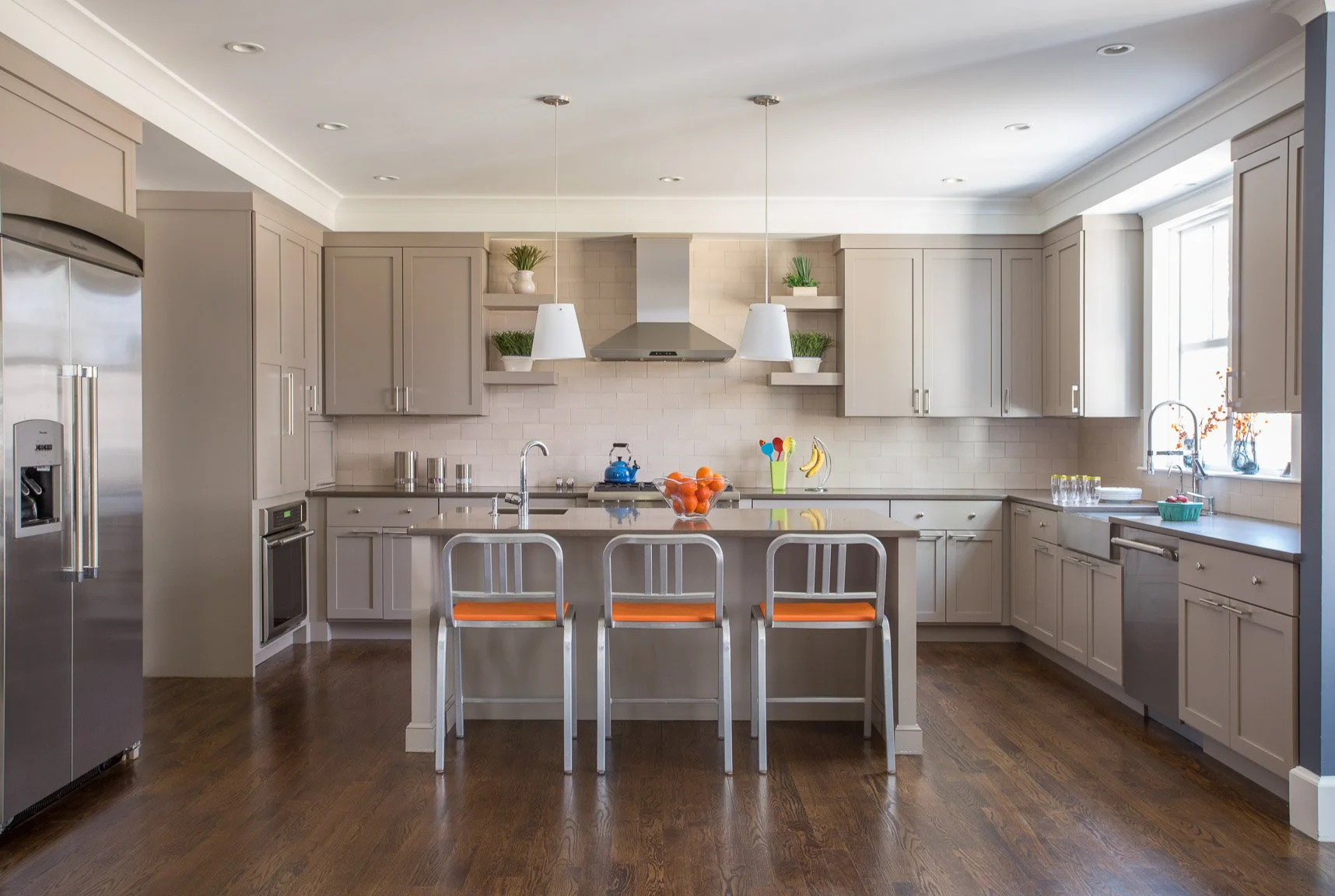 A modern kitchen with beige cabinets, stainless steel appliances, a kitchen island with three stools, and hardwood floors. Decor includes potted plants, a bowl of oranges, bananas, and colorful utensils.