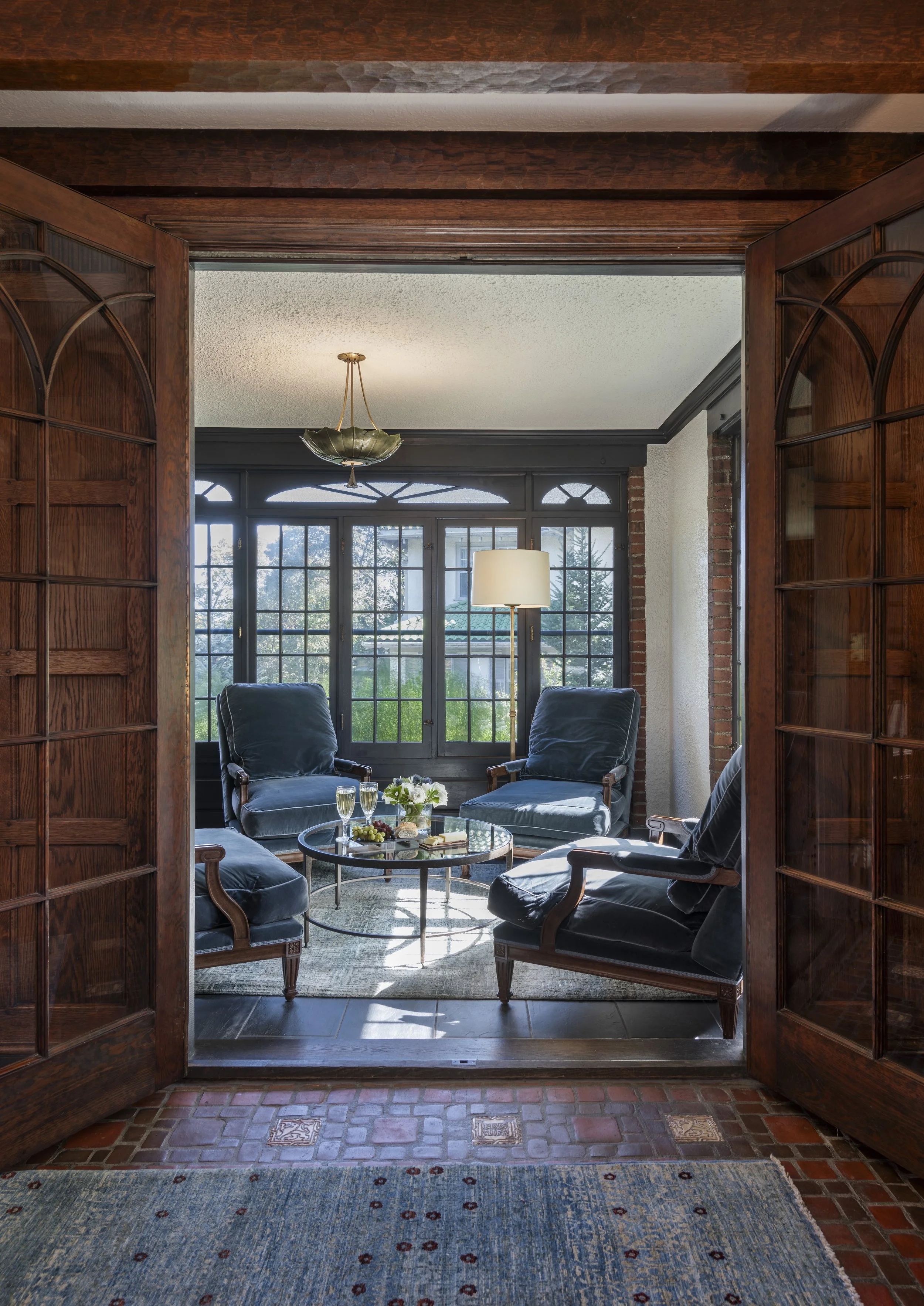 Tudor sitting room with velvet Louis XVI bergère chairs, brass leaf pendant, glass coffee table, and leaded glass windows.
