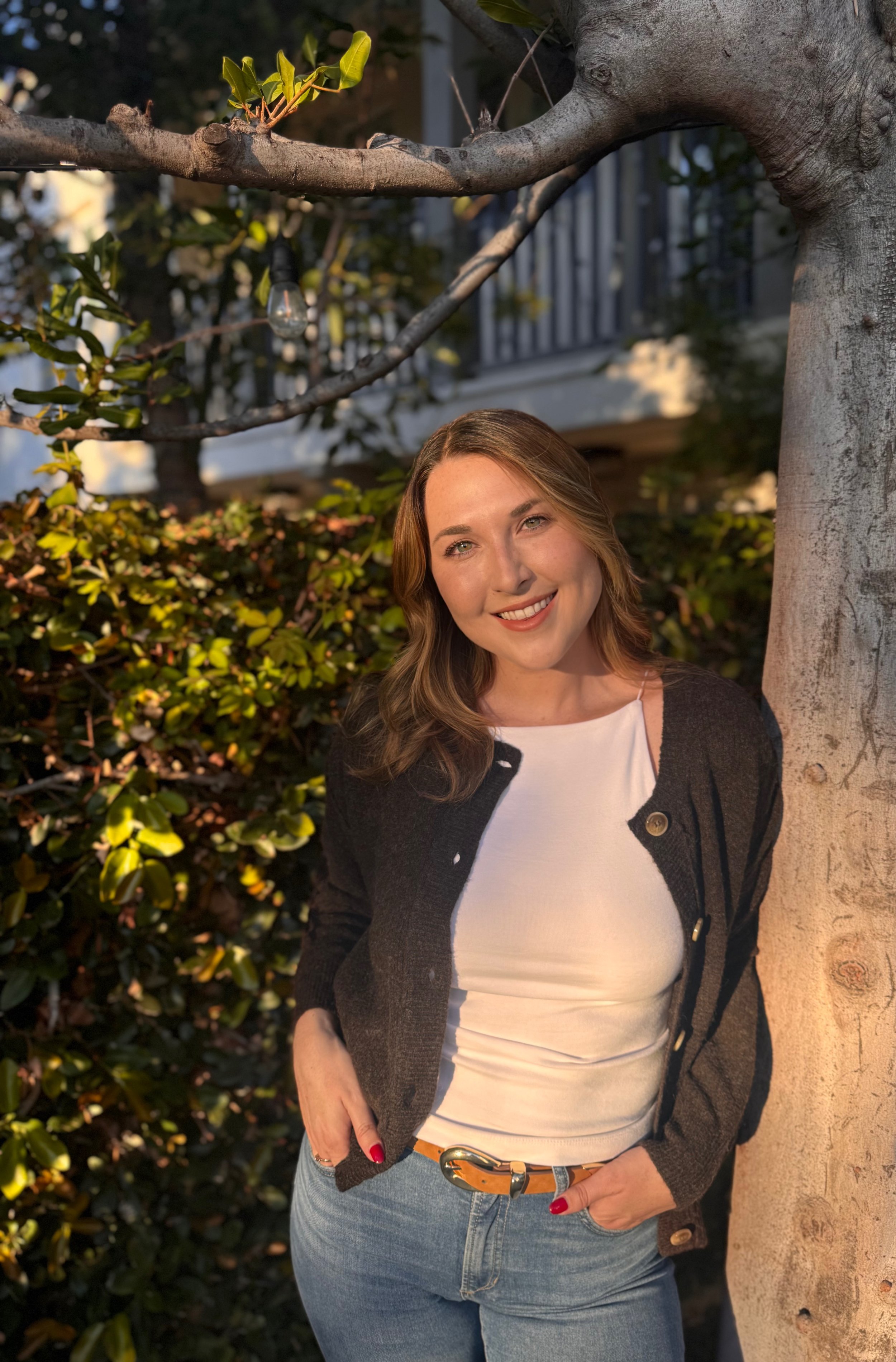 Brunette woman leaning against a tree in a backyard surrounded by greenery. She's dressed in a white top, with a brown cardigan and jeans with a belt.