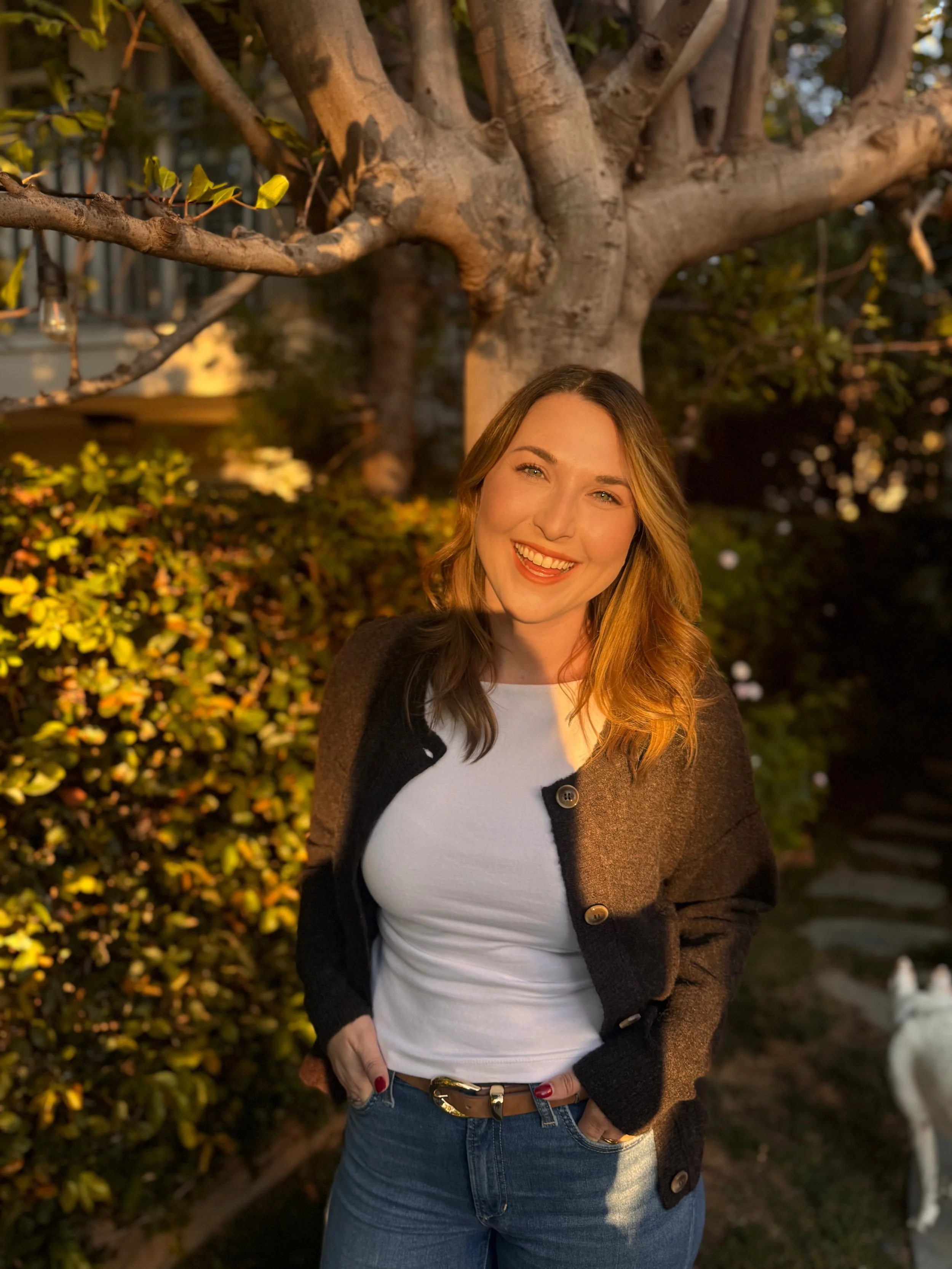 A young woman with light brown hair smiling outdoors at sunset, standing in front of a large tree and bushes.