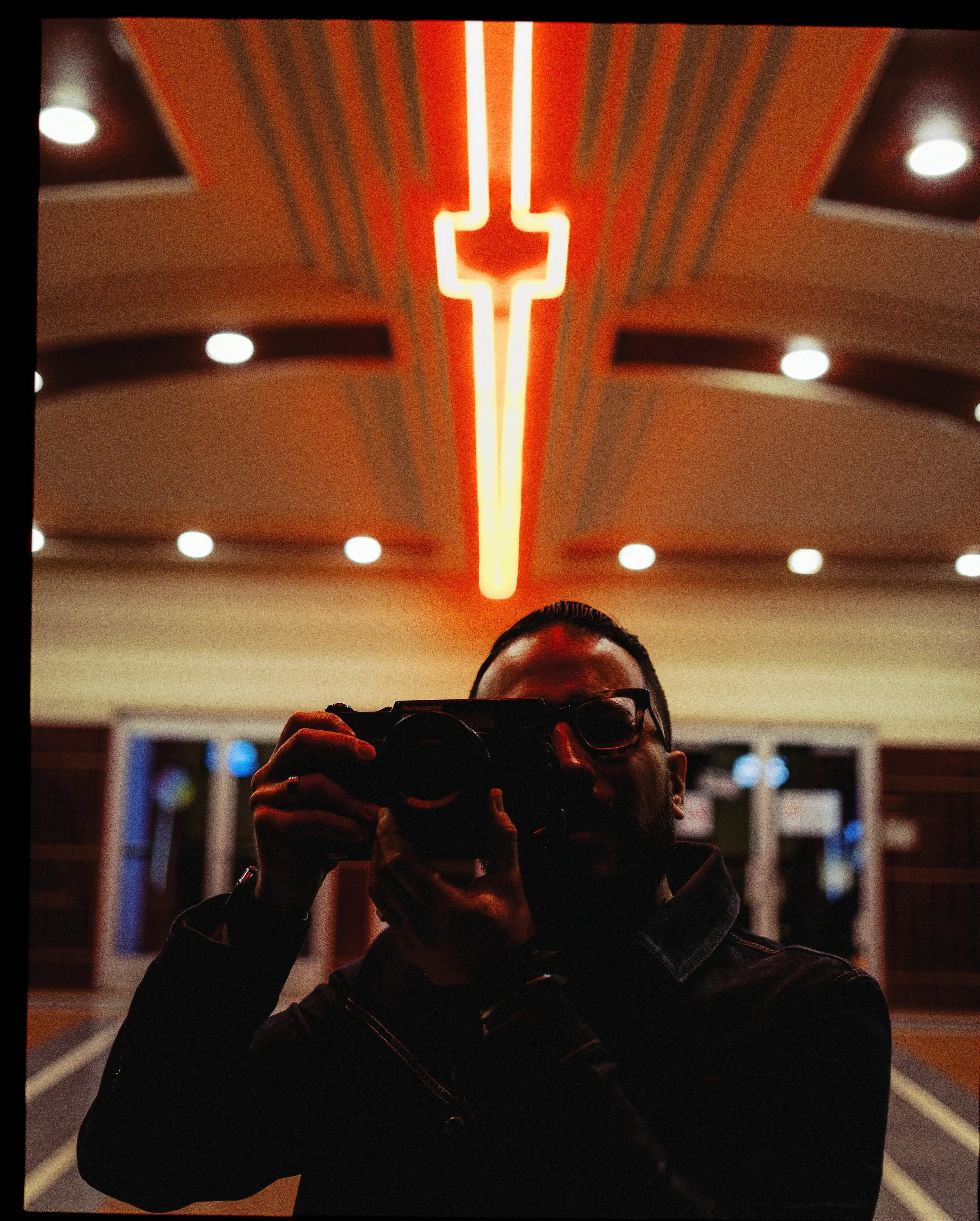 A person taking a photo with a medium format film camera, with a neon light in the shape of a cross above their head, outside an old school cinema with ceiling lights and double glass doors in the background.